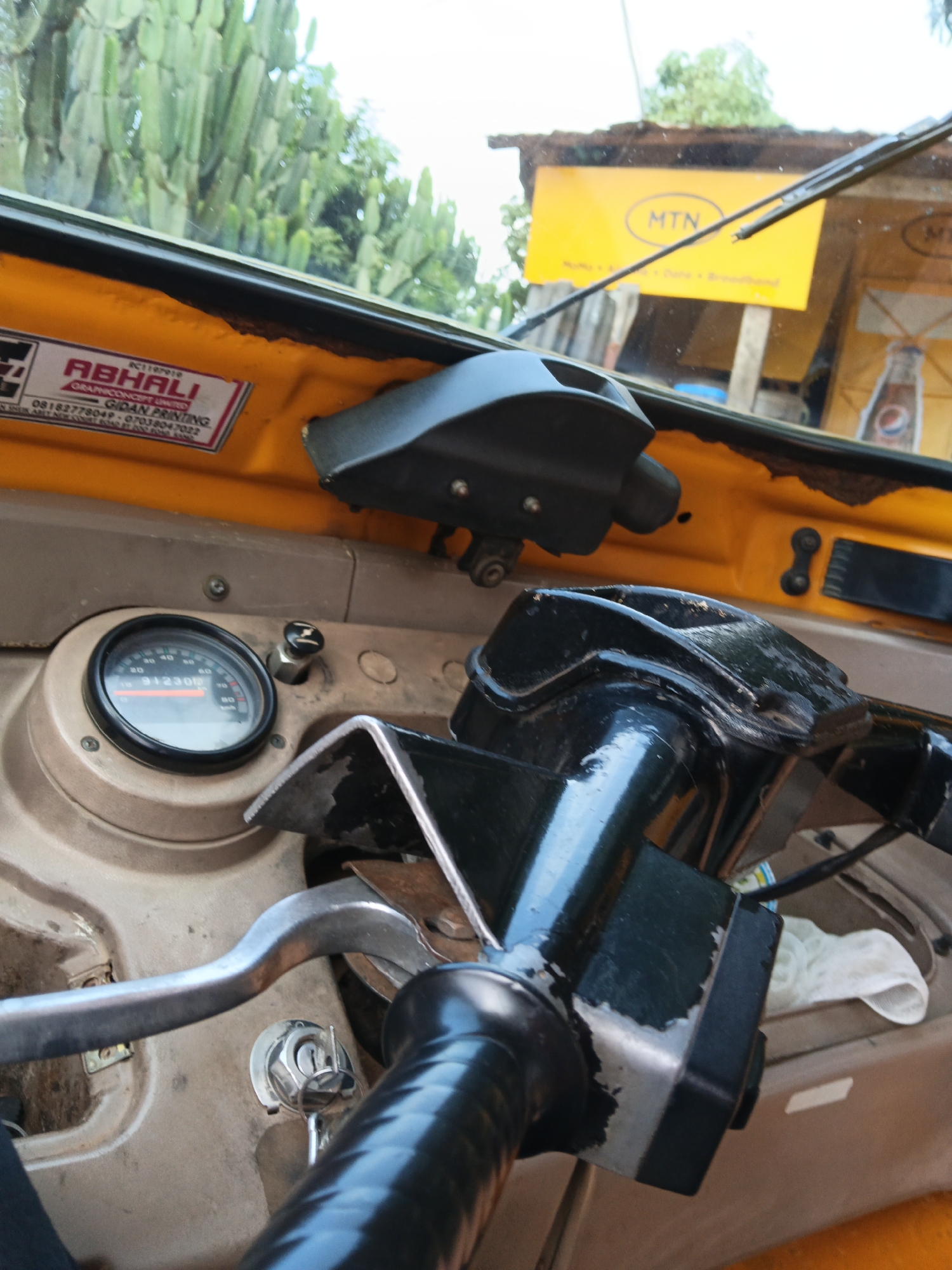 The image shows the interior of a yellow auto-rickshaw in Vom, Nigeria, with a partial view of the outside. The dashboard features a speedometer displaying 91230, a black steering column with a portion of the steering wheel visible, and a metal lever. A key is inserted in the ignition. Outside the vehicle, a large yellow sign with the MTN logo and text, along with a portion of a Coca-Cola advertisement, are visible, suggesting a roadside location. Green foliage, likely cactus plants, frames the top of the image, indicating an outdoor setting within or near the city.