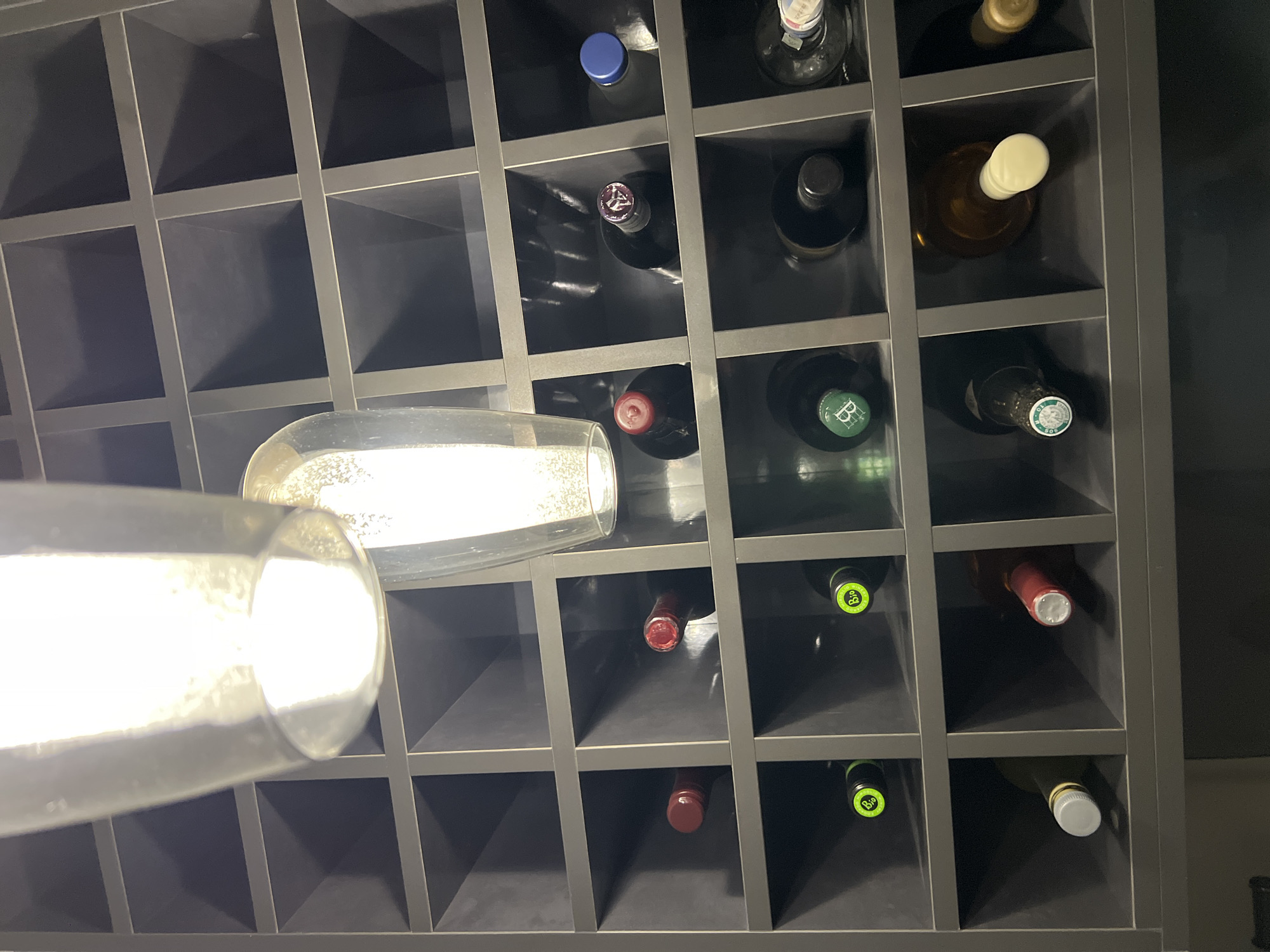 This media file captures a low-angle perspective of a dark-colored wine rack, filled with various bottles and featuring two prominent, brightly lit glasses in the foreground. The rack is constructed from a grid of horizontal and vertical dividers, creating square compartments that hold the bottles. Some compartments are empty, revealing the dark interior of the rack.
The bottles within the rack display a variety of caps and foil colors, including blue, purple with a stylized white symbol (possibly an 'Y' or 'V'), red with a white pattern, light green with a circular "B" logo (resembling "BIO"), and cream/white screw caps. One bottle in the upper right section has a label with faint Asian script, likely Japanese or Chinese characters. Many bottles are made of dark glass, suggesting they contain red wine, while others are lighter, possibly holding white wine or spirits.
The foreground is dominated by two highly illuminated, empty glass objects, likely wine or champagne glasses, which appear to be hanging or positioned in front of the rack. They are extremely bright, creating significant glare and reflections, obscuring parts of the rack behind them. The lighting in the image is uneven, with the glasses and certain bottle caps brightly lit by an unseen light source, while the rest of the rack and its contents remain in shadow.
There is no activity taking place, depicting a static arrangement of bottles and glasses. The setting is likely indoors. No specific time of day or weather can be discerned. The visible text includes the faint Asian characters on one bottle label and the "B" logo on the green caps. The location context is Burgas, Bulgaria.