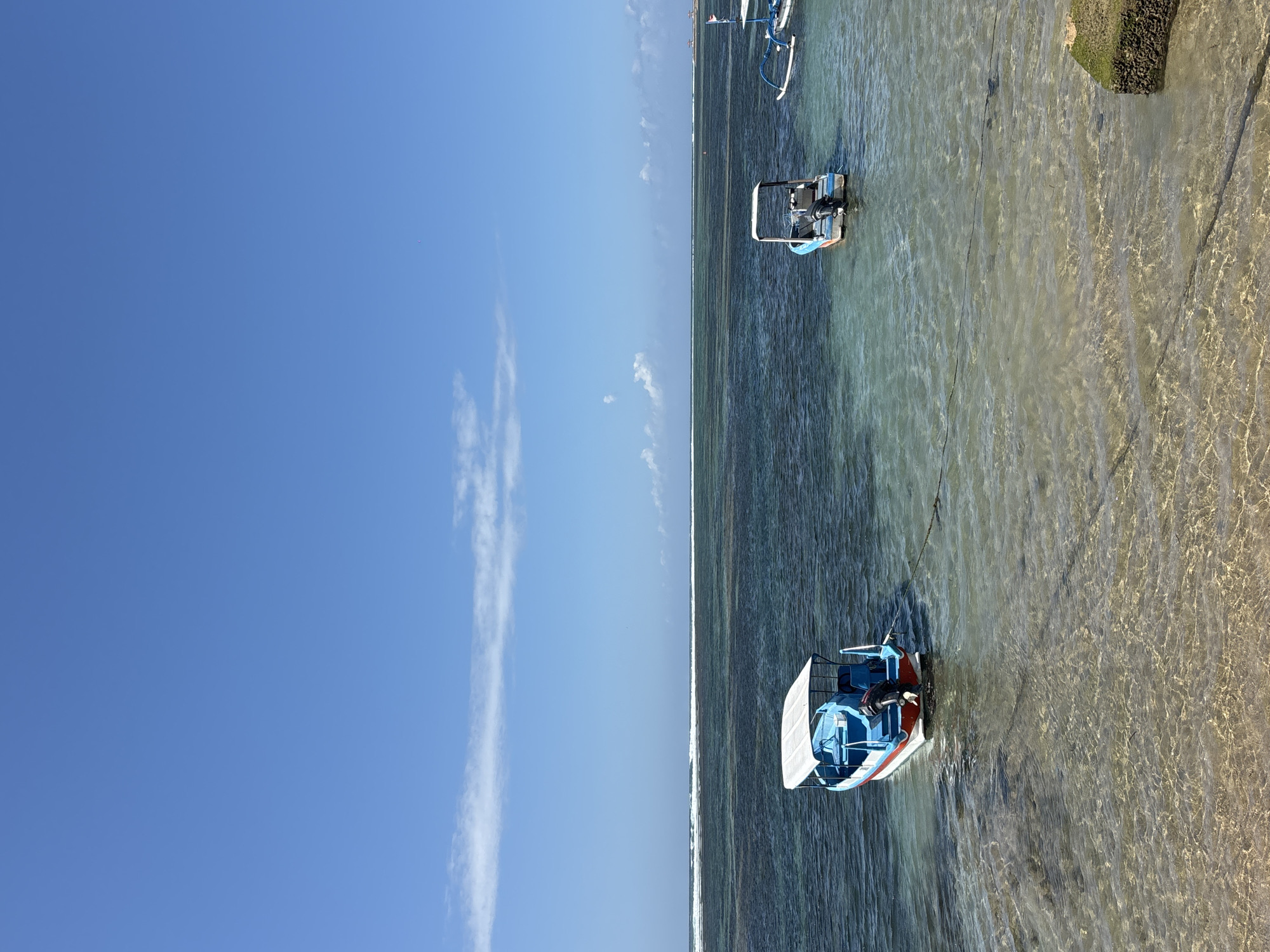 The image depicts a tranquil scene at a beach in Denpasar, Indonesia. The sky is a clear, vibrant blue with a few wispy white clouds scattered across it. The ocean water is a gradient of blues and greens, transitioning from shallow, crystal-clear shallows near the shore to darker, deeper waters further out. Gentle waves can be seen breaking in the distance along a reef.
In the foreground, the sandy seabed is visible through the transparent water. Two small motorboats are anchored in the water. The boat on the left is blue and white with a red stripe along its hull. It has a white canopy providing shade and an outboard motor at the stern. The boat on the right is similar in design, also blue and white with a white canopy and an outboard motor. A rope stretches across the water, anchoring the boats. In the far right of the image, a portion of a moss-covered stone structure is visible, suggesting the edge of a pier or seawall. The overall impression is one of peace and natural beauty.