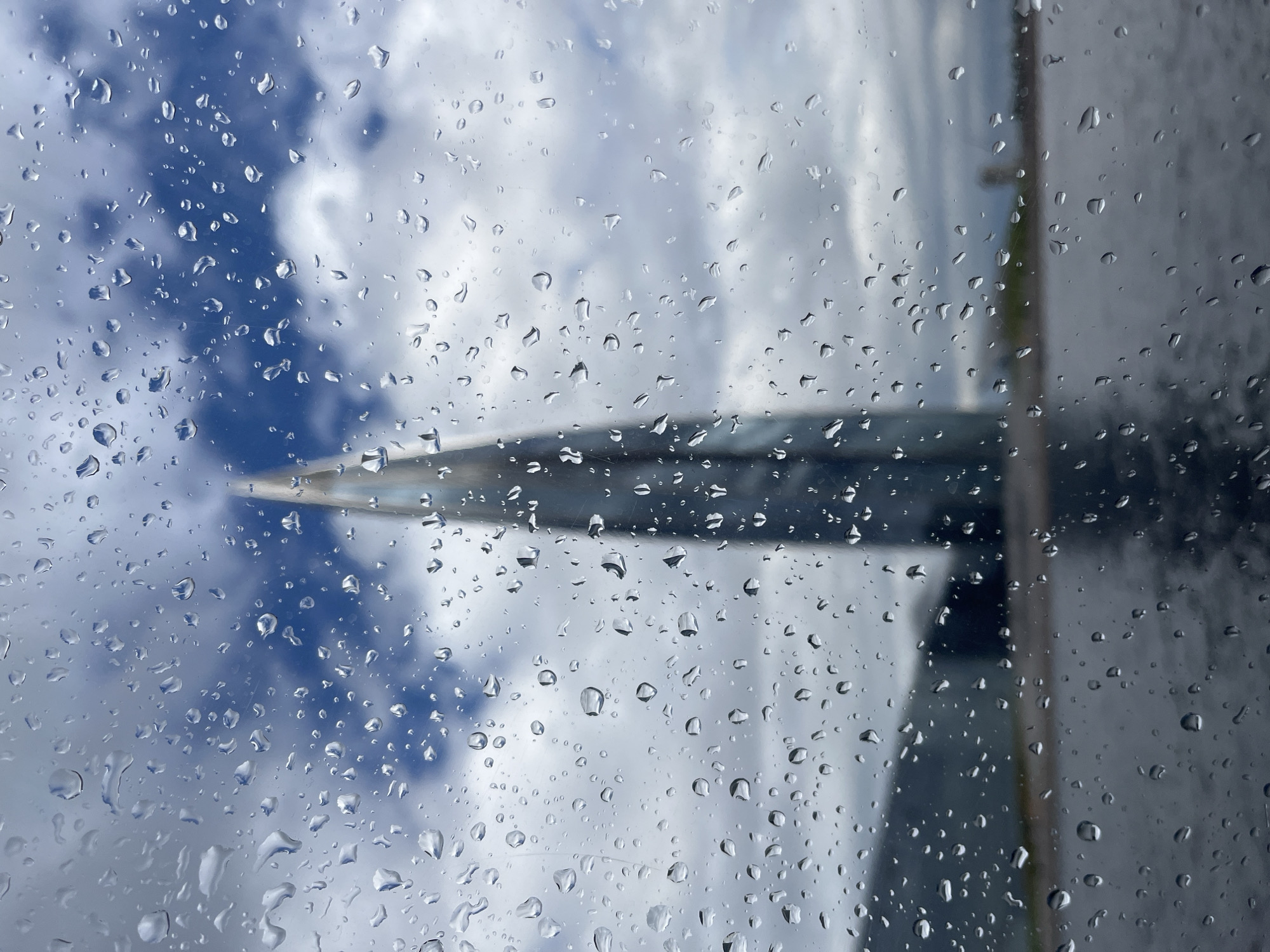 The image shows a close-up of a window covered in raindrops, with a blurred view of the sky and clouds behind it. The sky is a mix of blue and white, suggesting it is daytime. There appears to be a portion of an aircraft wing or fuselage visible through the window, also out of focus. The raindrops create a textured overlay, distorting the view of the sky and the aircraft component. The overall impression is one of being inside a moving vehicle, likely an airplane, during a rainy flight. No people, text, or specific emotions are evident. The location context of Saint Petersburg, Russia, suggests the flight may be in or out of that city.
