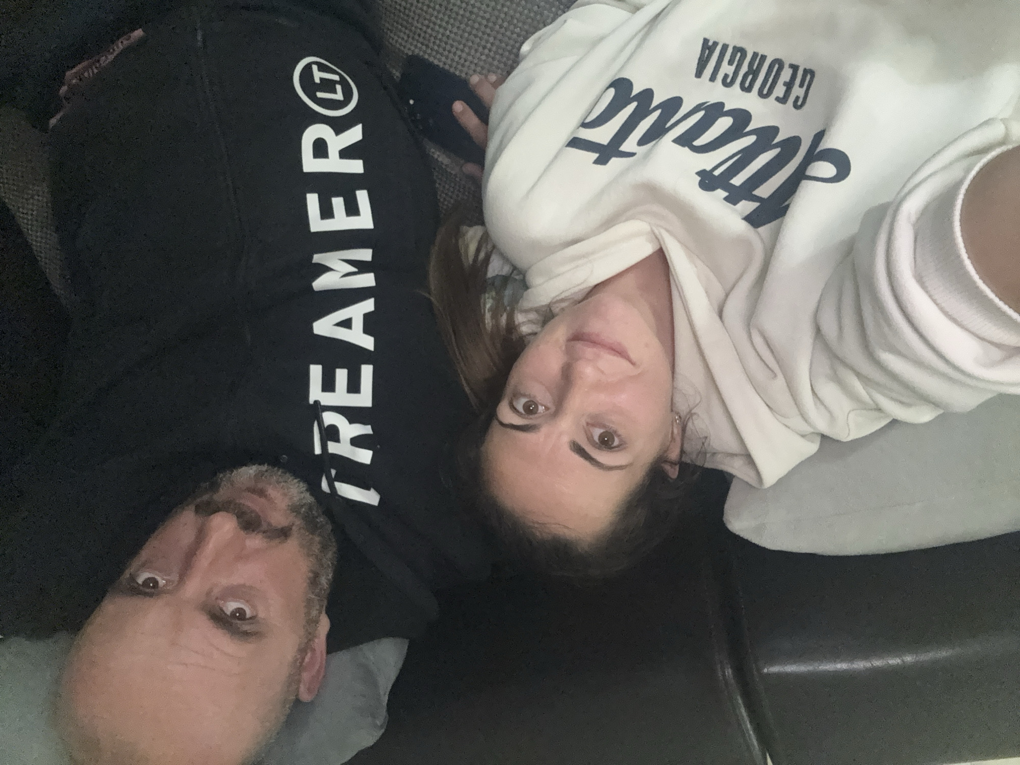 A photograph depicts a man and a woman lying down, presumably on a sofa. The man, positioned on the left, is wearing a black hoodie with the word "STREAMER" and the letters "LT" in a circle printed vertically on the sleeve. He has short, graying hair and a beard. The woman, on the right, is wearing a light-colored sweatshirt with the words "Atlanta, Georgia" printed on it. Their faces are turned towards the camera, suggesting they are taking a selfie. The background is somewhat blurry, showing a gray upholstered surface. The scene appears to be indoors. There is no information available to confirm that this photo was taken in Varna, Bulgaria.