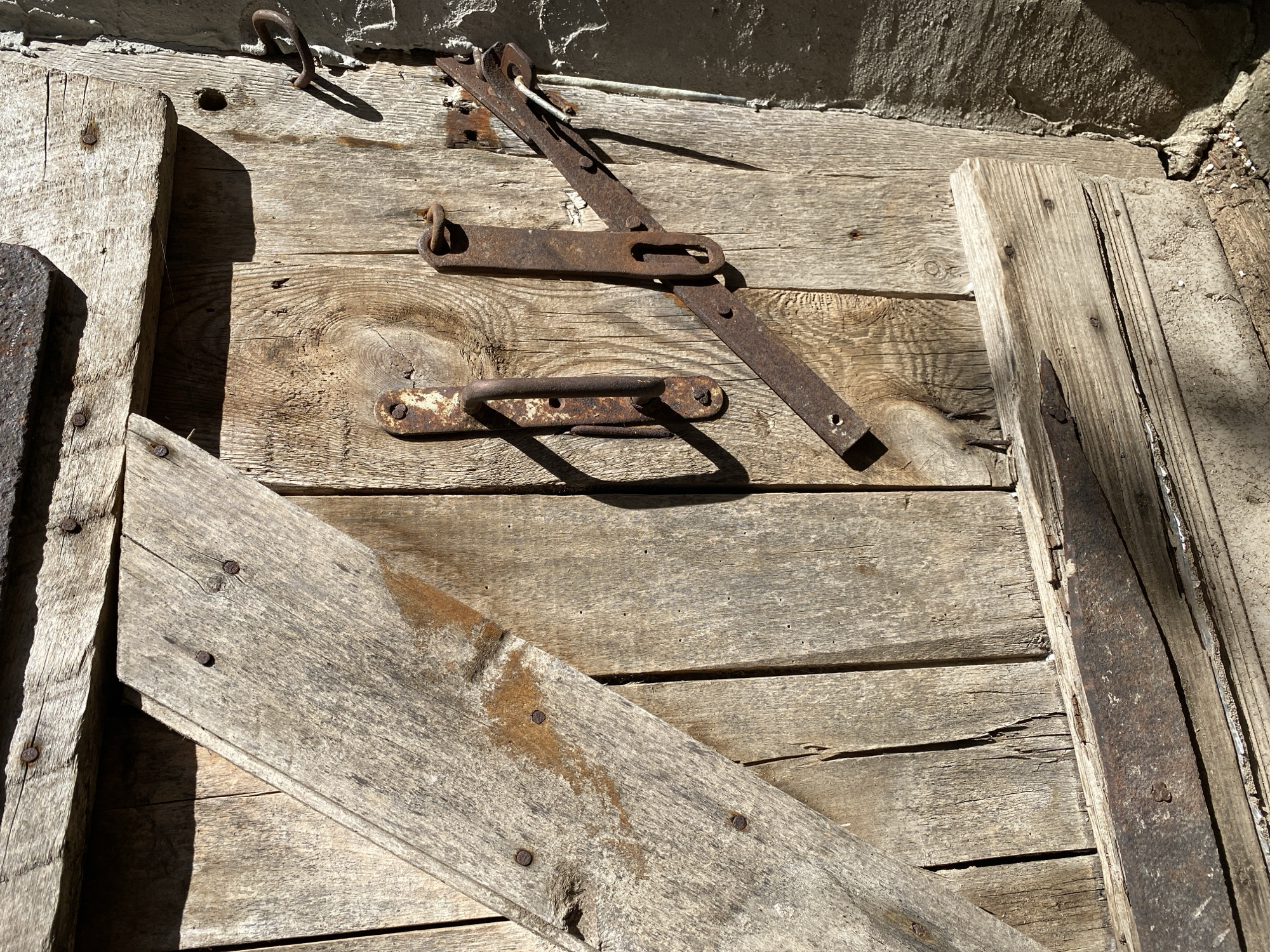 The image shows a close-up, detailed view of an old, weathered wooden door with rusty metal hardware. The door is constructed from vertical planks of wood, some of which are showing signs of age and wear, including cracks and splinters. A diagonal wooden brace reinforces the left side of the door, attached with several nails. The hardware consists of a worn, rusty door handle attached vertically to a plank, and a more complex locking mechanism. This lock includes a horizontal bar that swivels on a pivot, with a loop at one end that can engage with a staple or ring. A separate staple with a ring is also visible higher up on the door. The door is situated next to a rough, textured stone or stucco wall. The lighting suggests a bright, sunny day, casting distinct shadows. The specific location in Vitsebsk, Belarus is not discernible from the visual information alone, as the scene depicts a generic architectural element common to older structures. No people or animals are present in the image, nor are there any discernible actions or interactions occurring.