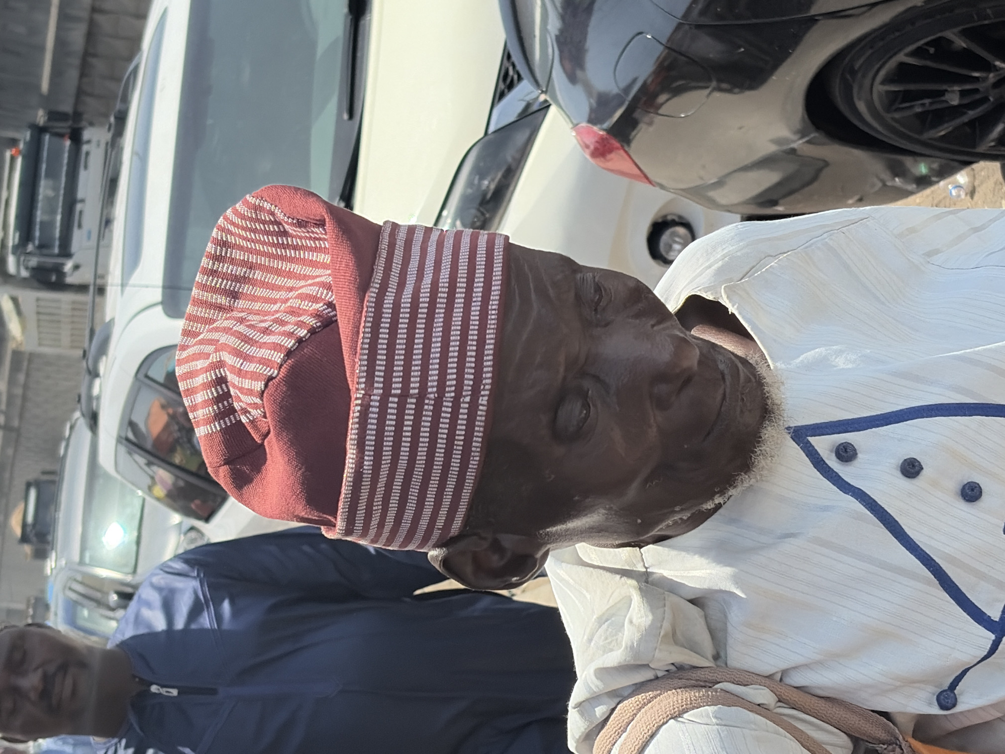 An elderly man wearing a red and white striped hat and a white shirt with blue accents is the primary subject of the photograph. He has dark skin and a white beard. The man appears to be standing outdoors near several vehicles, including a black car on the right edge of the frame. The background includes a line of white vans. Another person wearing a blue jacket is partially visible in the lower left corner. The setting appears to be a sunny outdoor location in Lekki, Nigeria, possibly a parking lot or street.