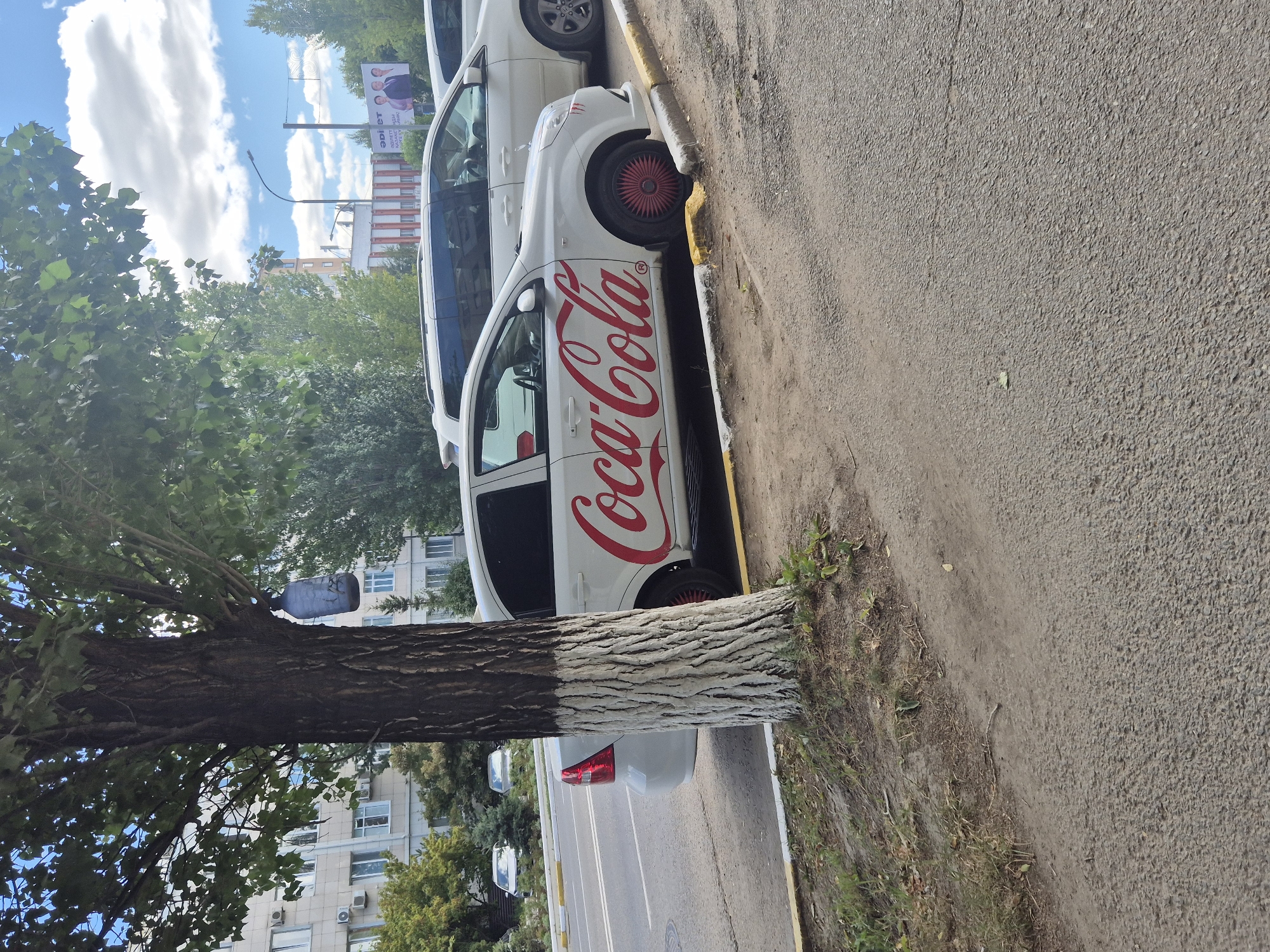 A white Chevrolet Aveo with a large "Coca-Cola" logo on its side is parked on the right side of a paved road in Kokshetau, Kazakhstan. The car has red hubcaps. A tree with a whitewashed trunk is to the left of the car, obscuring part of its front. In the background, there are buildings, including an apartment complex and a building with a striped facade. A billboard with an image of three men is visible in the distance. Other vehicles, including a white minivan and two silver sedans, are also present in the scene. The sky is partly cloudy.