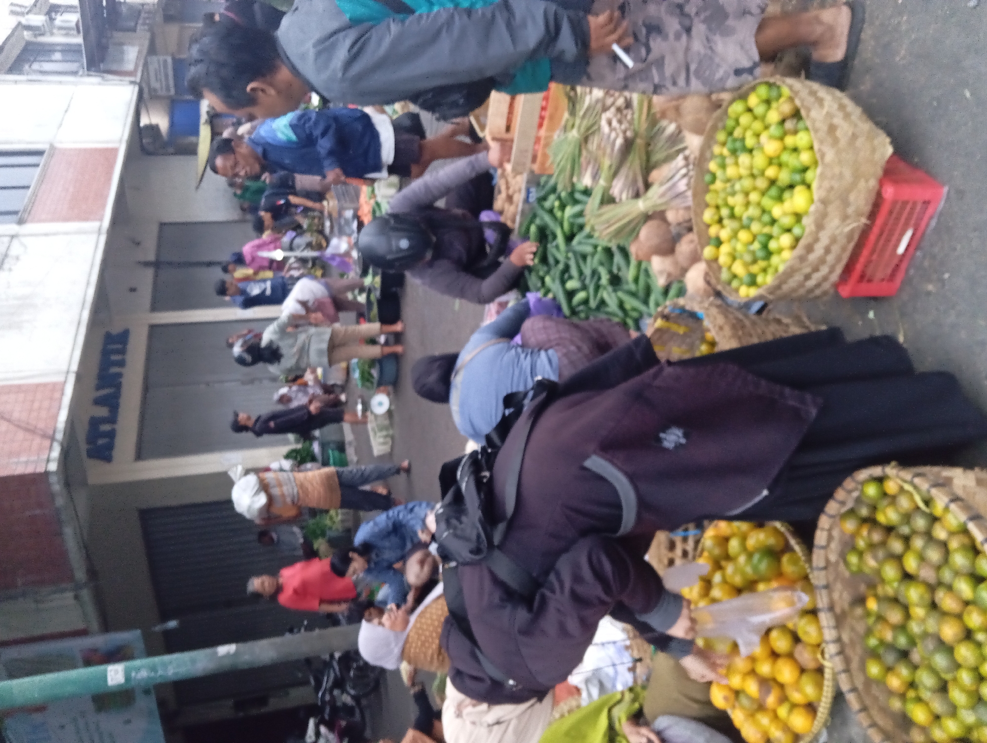 In a street market in Yogyakarta, Indonesia, a bustling scene unfolds. People are seen engaged in various activities, from selling to shopping, amidst a vibrant display of produce. In the foreground, two large woven baskets are filled with fresh citrus fruits, one containing oranges and the other a mix of green and yellow limes. Nearby, a woman in a dark hijab and robe is reaching into one of the baskets, seemingly selecting fruits.
Behind the fruit displays, a variety of vegetables, including cucumbers and bundles of herbs, are laid out. Several individuals are interacting with the produce, some appearing to be vendors organizing their goods and others customers browsing. The market is situated in front of a building with signage that reads "ATLANTIK". The overall atmosphere suggests a lively commercial hub, typical of a local market in a city.