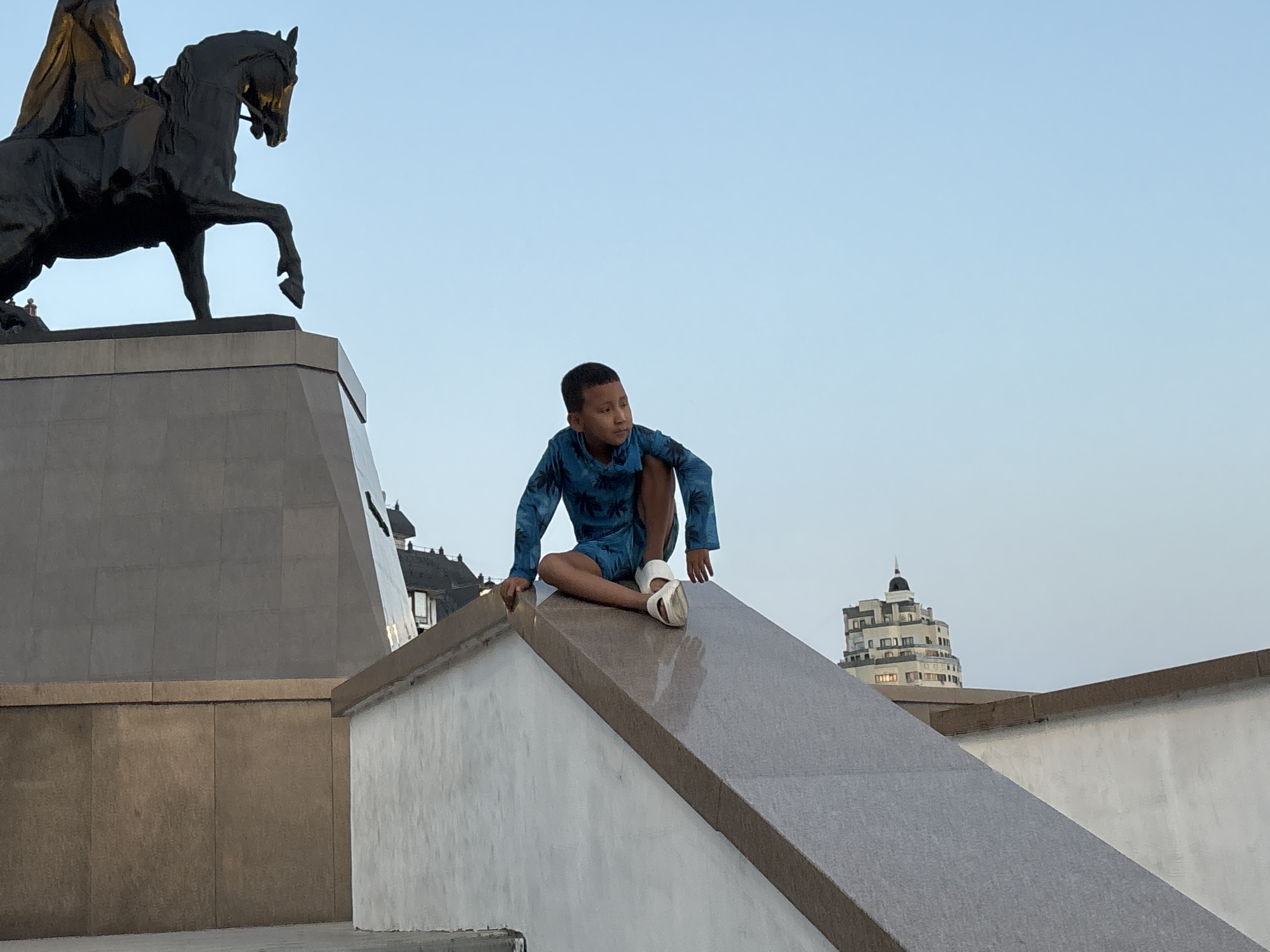 A young boy in a blue patterned long-sleeved shirt and shorts, wearing white sandals, is crouched on a slanted stone structure in front of a monument. The monument features a dark bronze statue of a person on horseback. The background shows a clear, pale blue sky with a tall, light-colored building in the distance. The scene appears to be outdoors in an urban setting, likely a public plaza or park.