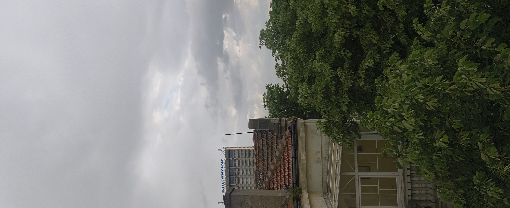 The image shows a view of Varna, Bulgaria, taken at an upward angle. The sky is overcast with a mix of gray and white clouds. A building with a terracotta tile roof is visible in the lower part of the frame. Next to the roof is a facade featuring windows. A tall, modern building with the sign "HOTEL CHERNO MORE" is seen in the background. Green trees fill the right side of the image, partially obscuring the building facade.