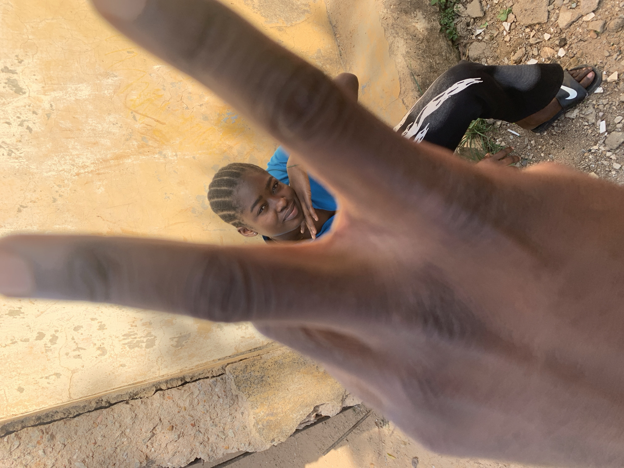 The image shows a young woman in Rafin Par, Nigeria, making a peace sign with her hand. She is wearing a blue shirt and has braided hair. Her right hand is placed on her left arm. She is looking towards the camera. She is next to a yellow building with damaged or crumbling concrete along the base. The ground surrounding the building is dirt with some rocks and small patches of vegetation. A person wearing black leggings with white accents and black Nike sandals is also partially visible.