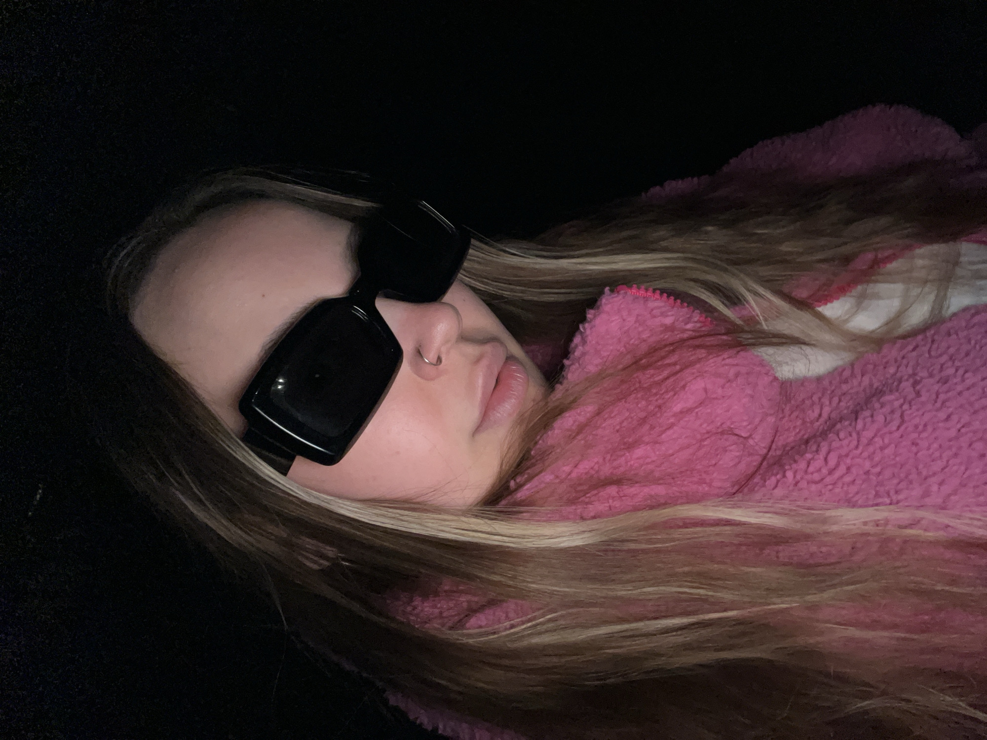 A young woman wearing black sunglasses and a nose ring lies down, partially obscured by a pink fuzzy jacket. The background is dark, suggesting a poorly lit indoor setting.