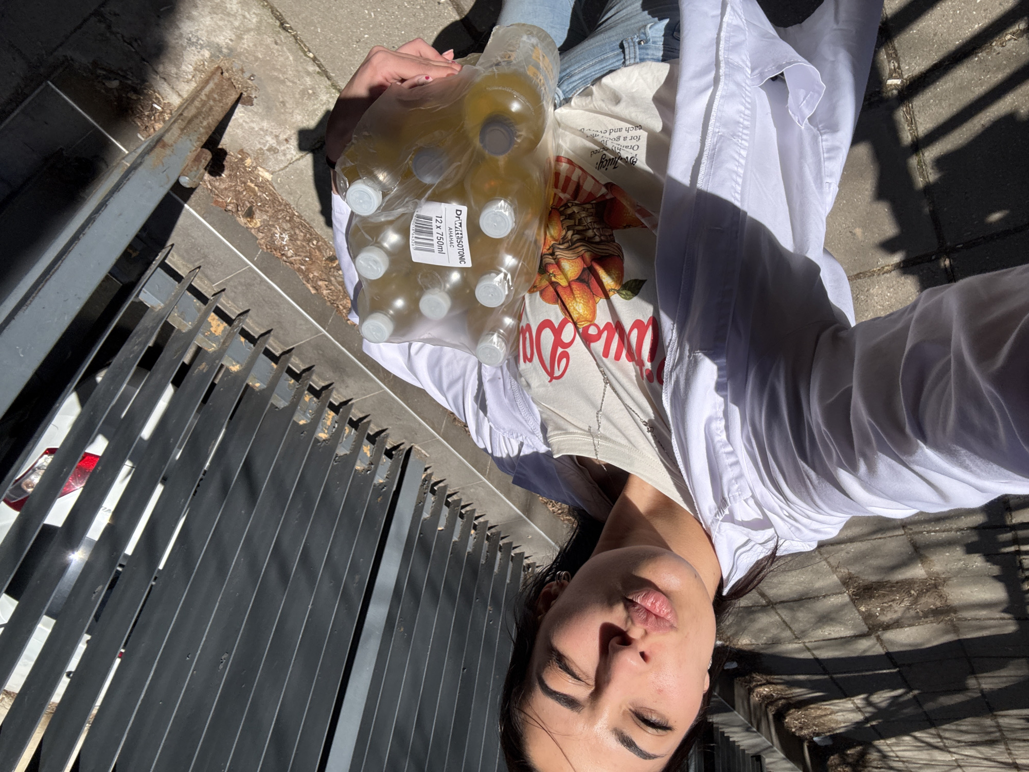 The image shows a person lying on a paved surface, likely a sidewalk, next to a metal grate. The person is holding a plastic-wrapped pack of twelve 750ml bottles of DrWISOTONIC AHAHAC. They are wearing jeans, a graphic t-shirt with an orange motif and the word "Juicy", and a white shirt. The scene appears to be outdoors in a sunny location. The city of Sofia, Bulgaria, is not discernible from the image alone.