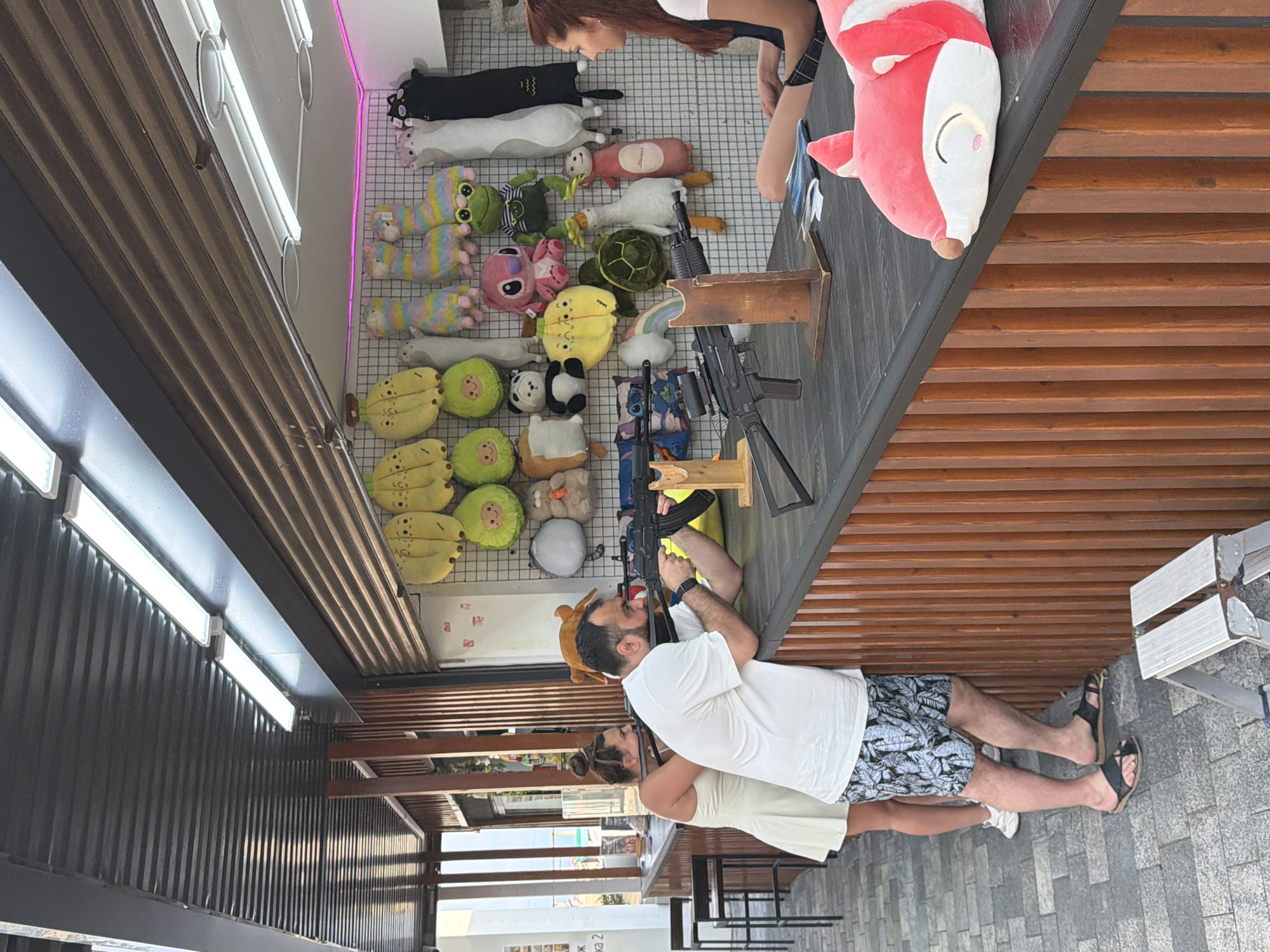 The image depicts an outdoor shooting gallery attraction, likely located in Sochi, Russia, given the presence of beachgoers and the general ambiance. In the foreground, a man in a white t-shirt and patterned shorts is aiming a replica rifle at a target, with a woman in a white dress standing behind him, also looking in the direction of the target. The shooting gallery counter is stocked with various plush toys, including bananas, avocados, cats, and other cartoon characters, which likely serve as prizes. The counter itself is made of dark wood and is lined with vertical wooden planks. To the right, a woman with red hair is seated, observing the scene. The background shows a corrugated metal roof and fluorescent lighting, suggesting an open-air market or amusement area. The ground is paved with grey interlocking stones.