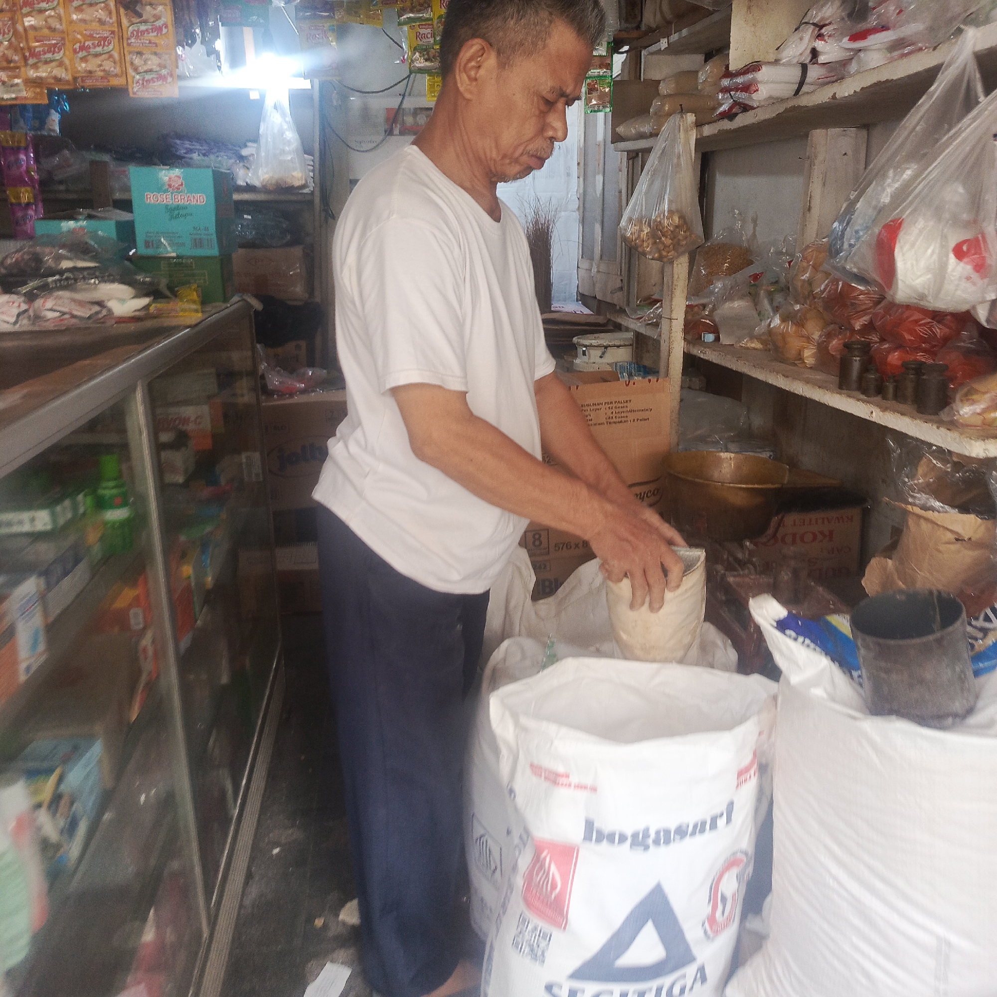 A middle-aged Indonesian man with graying hair and a light complexion is standing in what appears to be a small grocery store or market stall. He is wearing a plain white t-shirt and dark blue trousers. The man is actively engaged in scooping a light-colored powdery substance from a small white bucket into a large white sack with red and blue lettering that reads "bogagasi" and "SEGITIGA". His posture suggests he is focused on this task.
The environment is cluttered with various goods commonly found in such establishments. Shelves are visible in the background, stocked with packaged food items, including what look like snack packets hanging from above, and boxes of products. A glass display case is positioned to the left, showcasing more items. Various sacks, bags, and containers are present throughout the scene, some containing goods and others empty. A set of old-fashioned brass weights is visible on a shelf. The lighting appears to be from overhead fixtures, creating some bright spots and shadows. The overall impression is of a busy, functional retail space.
The provided crops indicate that this scene is located in South Jakarta, Indonesia, specifically in the Tebet area, likely within a neighborhood market or a small independent shop.