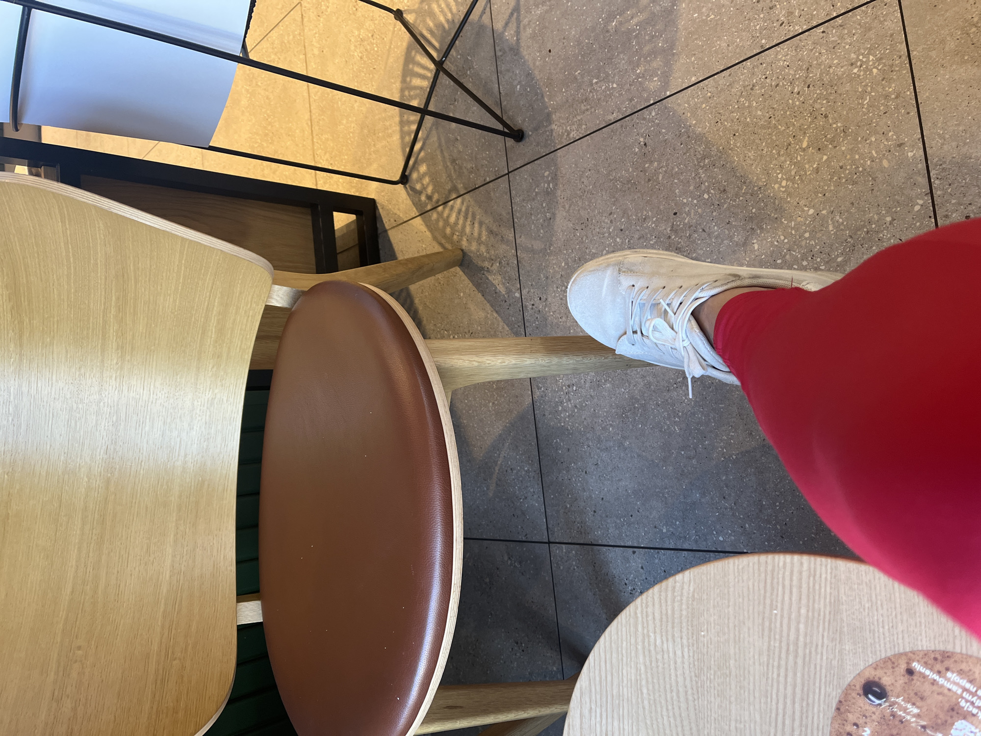 The image shows a person's legs in red leggings and white sneakers, with their right foot resting on a chair. The scene appears to be indoors, possibly a cafe or restaurant, with modern wooden chairs and tables. The floor is tiled with a speckled gray pattern. A metal chair with a geometric design casts a shadow on the floor. There are also glimpses of other furniture, including a light-colored wooden chair and a round table with some writing on a coaster. The lighting suggests it could be daytime, possibly late afternoon given the shadows. The overall impression is of a casual, contemporary setting.