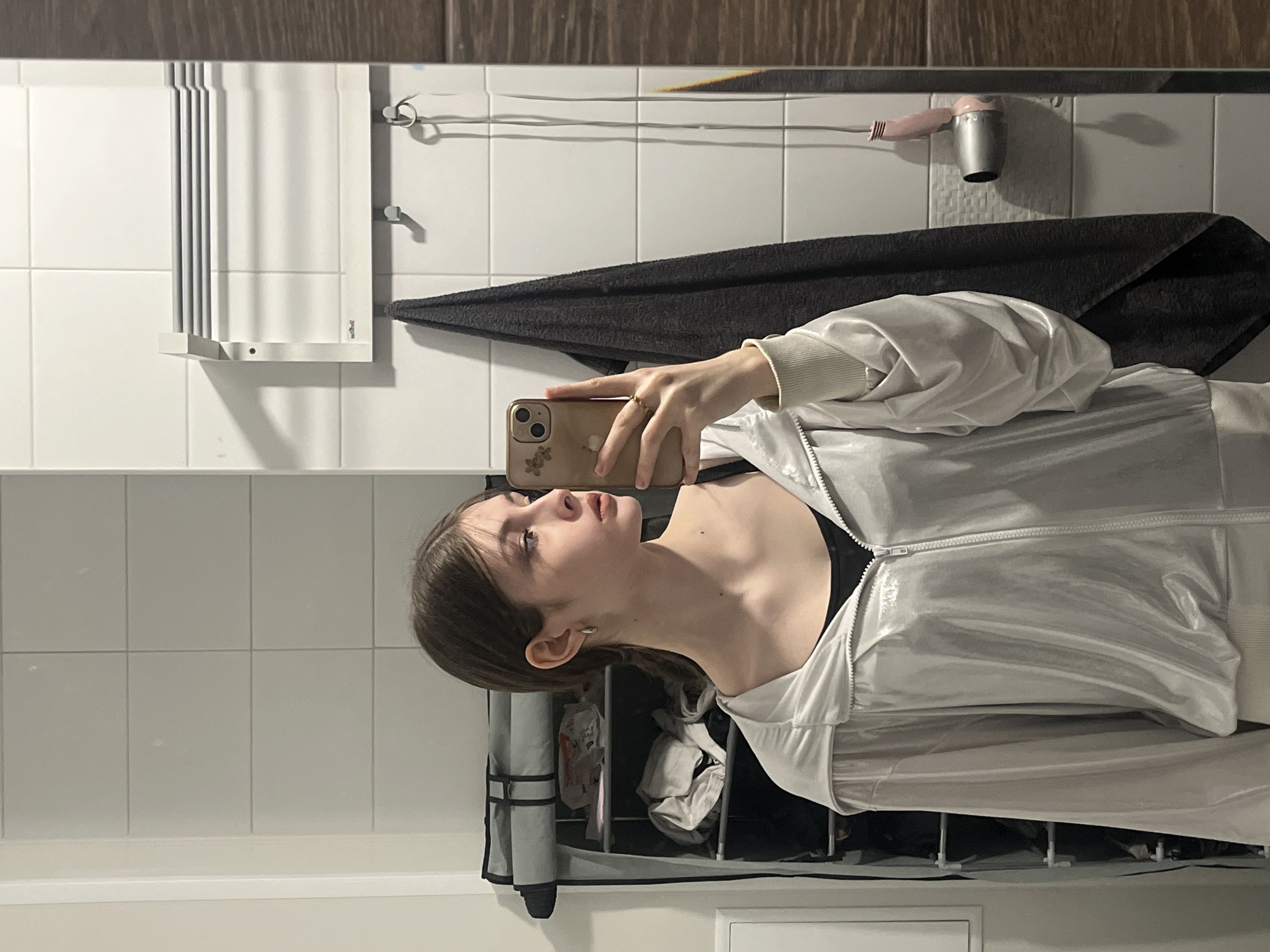 A young woman is captured in a mirror selfie taken in what appears to be a bathroom. She is positioned facing to her left (viewer's right) with her head tilted upwards, revealing her neck and chin. Her medium-length brown hair is swept back, and she has a neutral or slightly contemplative expression. She wears a light-colored, possibly silver or metallic, zippered jacket or tracksuit top over a black garment. A small stud earring is visible in her left ear.
She holds a gold or tan-colored smartphone with her right hand, its camera visible on the back, along with a floral design detail on its case. A ring is worn on a finger of the hand holding the phone.
The background shows a tiled wall with white rectangular tiles and light grey grout. Along the top of the wall, there's a dark wooden-textured border. Various bathroom fixtures are visible: a white, multi-bar towel warmer or drying rack is on the left. Below it, a dark grey towel hangs from a hook. Further to the right, a small, pink and silver hairdryer is mounted on the wall. Below the woman's torso, a multi-compartment fabric organizer, possibly for storage or laundry, is visible, containing miscellaneous items. The bottom of the image shows a white border, likely the top edge of a counter or bath fixture. No discernible text is clearly visible.