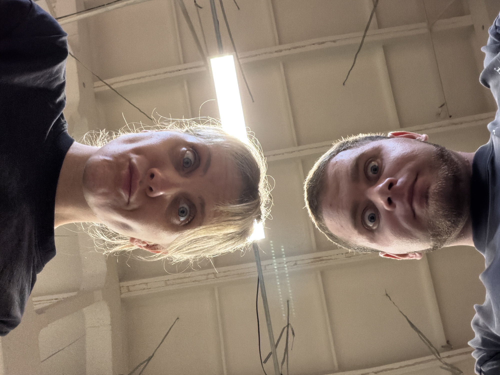 Two people, a female on the left and a male on the right, are looking downwards at the camera. The female has blonde hair and blue eyes. The male has short brown hair and a beard. They are in an industrial-looking space with a white ceiling and fluorescent lighting. The exact location in the city of Chaikovsky, Russia, is unknown based on the image alone.