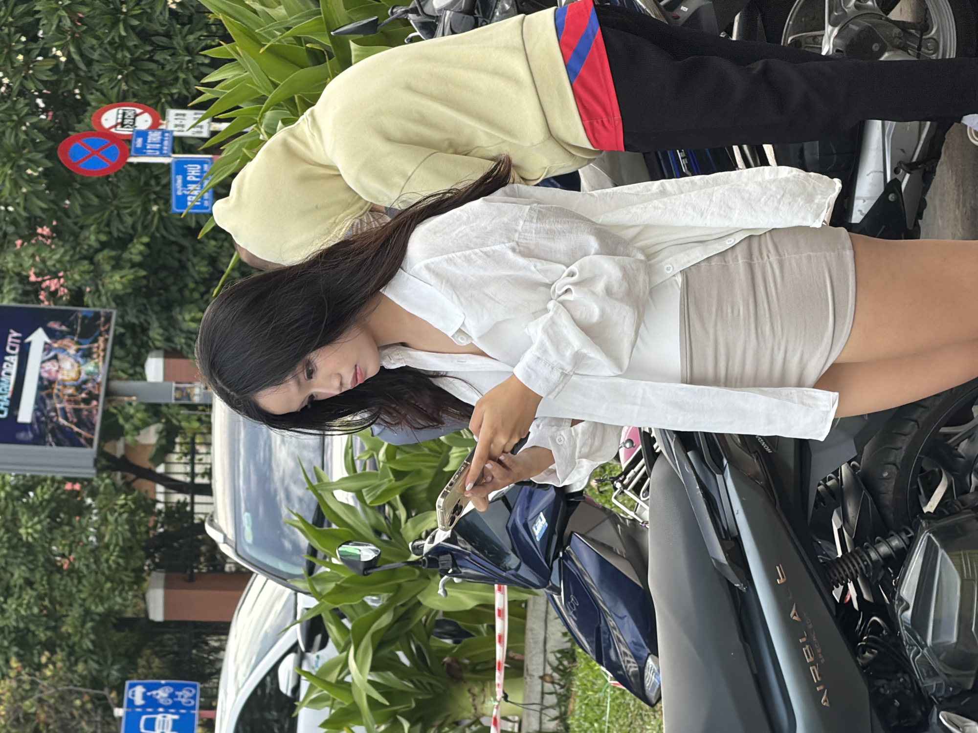 In Nha Trang, Vietnam, a young woman with long dark hair, wearing a white button-down shirt and a beige skirt, stands outdoors and looks down at her phone. She is positioned in front of a dark blue scooter. To her right, a person wearing a light yellow hooded sweatshirt with a red and blue striped shirt underneath, and black pants, is partially visible, also looking downwards. In the background, there are green plants, a white car, a gray SUV, and various traffic signs, including one with a blue background and white icons of a car, bicycle, and motorcycle, and another red and blue circular sign. The ground is paved and covered with some grass.