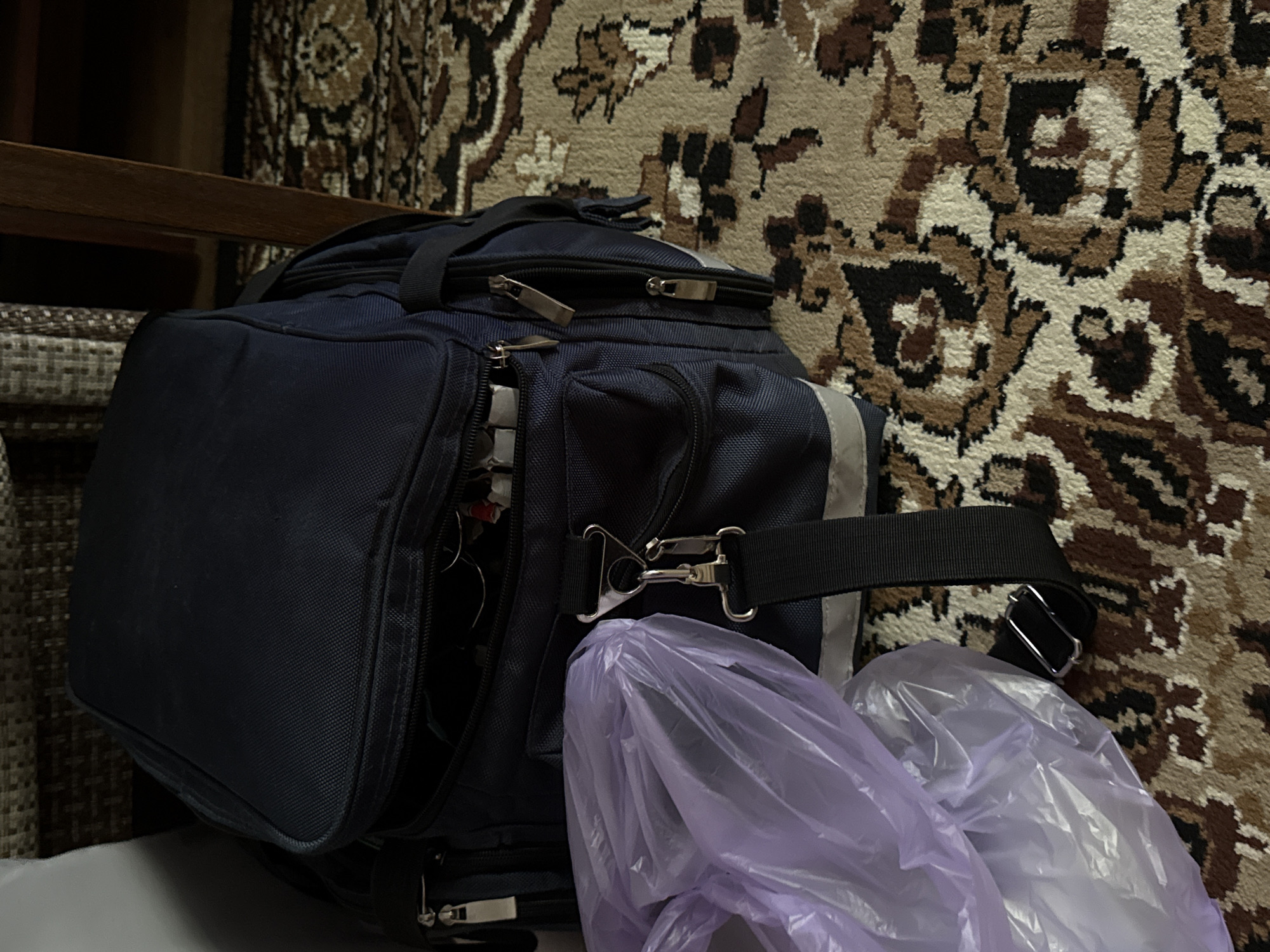 A dark blue backpack is positioned on a patterned carpet. A purple plastic bag is partially covering the front of the backpack, obscuring some of its details. The backpack has multiple zippers and straps visible. In the background, a dark wooden furniture leg and a portion of a patterned textile are discernible. The lighting suggests an indoor environment.