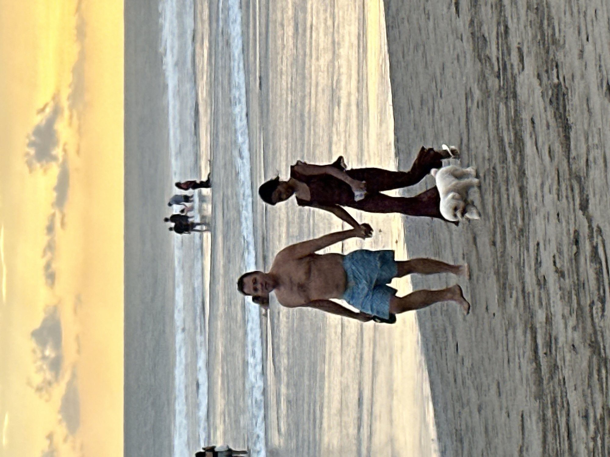 A couple walks hand-in-hand on a beach with a small white dog in North Kuta, Indonesia. The man, bare-chested and wearing blue swim trunks, walks slightly ahead of the woman, who is dressed in a dark red outfit. The dog, a fluffy white breed, walks to the woman's right. In the background, the ocean is visible with waves crashing on the shore and several other people are in the water and on the beach. The sky is a soft yellow and orange, indicating either sunrise or sunset, with scattered clouds. The sand is wet and reflects the light from the sky.