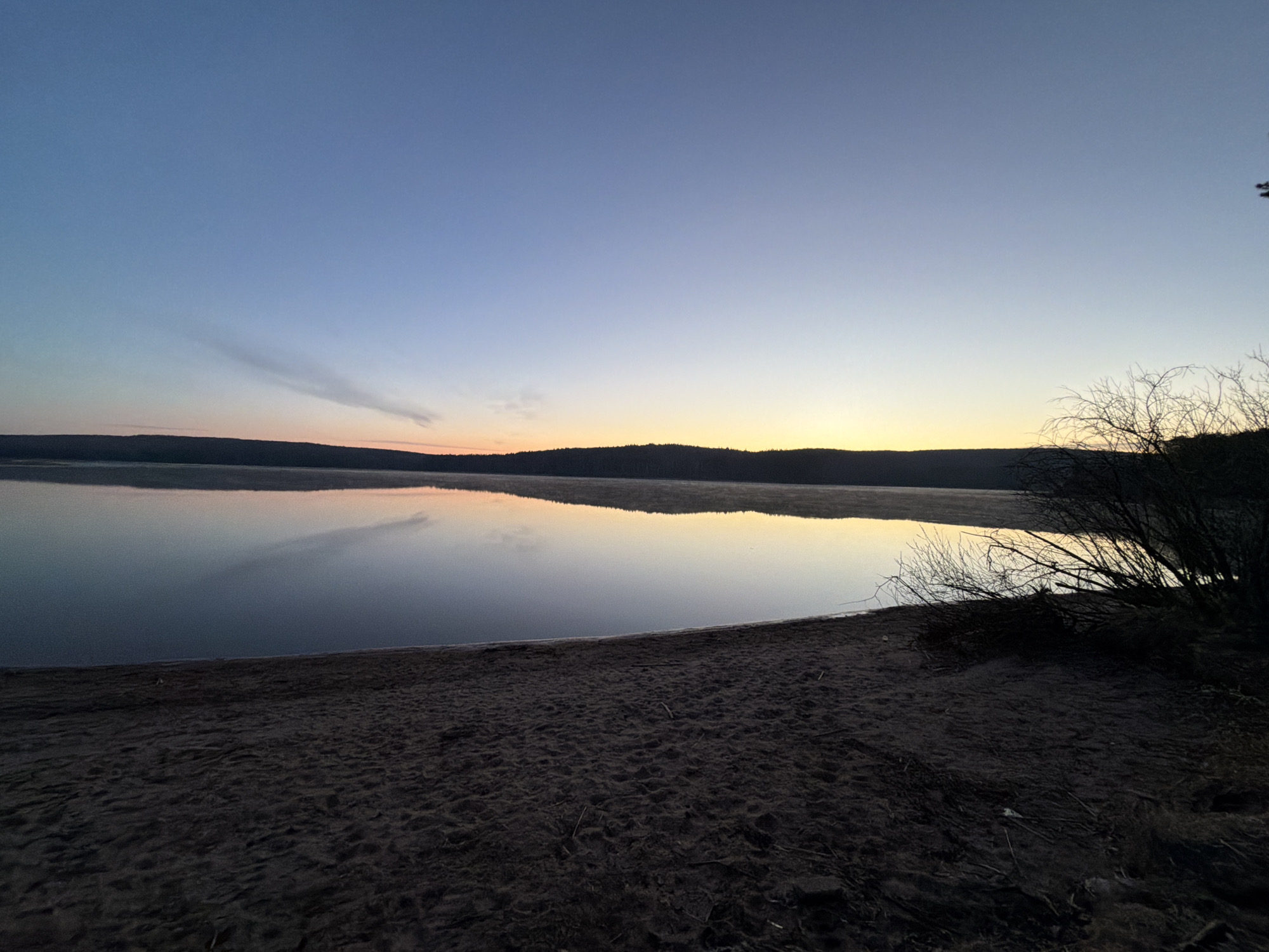 The image captures a serene natural landscape at either dawn or dusk in Chaykovsky, Russia. The scene is dominated by a calm, reflective body of water, likely a lake, which occupies the midground and reflects the soft colors of the sky.
In the foreground, a dark, sandy or dirt beach curves along the bottom, showing subtle textures and scattered small debris like twigs and leaves. To the right, a cluster of dark, silhouetted bare branches from bushes or small trees extends into the frame, partially obscuring the water's edge.
The horizon is marked by a dark, distant forested shoreline. Above this, the sky transitions smoothly from a soft orange and yellow glow where the sun is rising or setting, fading into a clear, deep blue overhead. A distinct, elongated wisp of cloud stretches across the upper left portion of the sky. The water's surface is remarkably still, creating clear reflections of the sky and the distant trees, with a faint suggestion of mist or haze hovering over the water further out.
No people are visible, and there is no apparent activity taking place. The overall impression is one of tranquility and natural beauty. No visible text is present in the image.