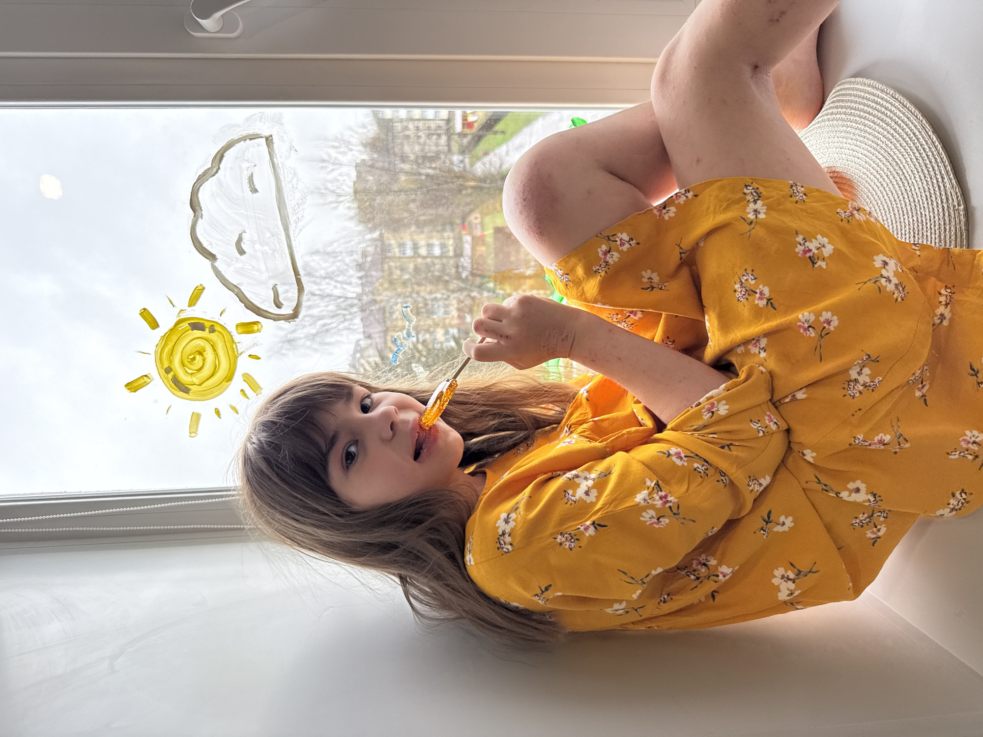 A young girl is sitting in front of a window, possibly in an apartment or home in the city of Rusanovo, Russia. She is wearing a mustard yellow dress with small floral prints of white, pink, and green. She is holding a lollipop in her hand and placing it in her mouth.
On the window, there are drawings of a yellow sun and a white cloud, made with some kind of washable paint or marker. Outside the window, there's a view of buildings and bare trees. The girl is sitting with her legs crossed and resting on a white surface, and a straw hat sits next to her. The background is out of focus.