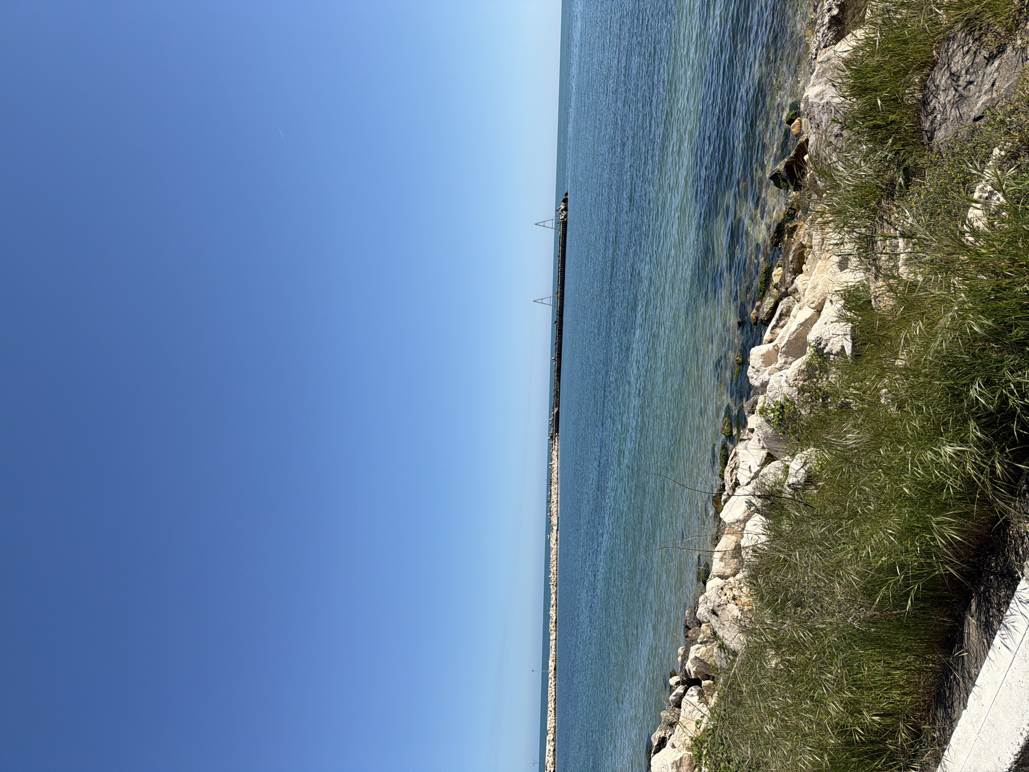 A daytime view of the Black Sea from Varna, Bulgaria. The sky is clear and blue. The water is calm with a pier extending into the sea. The shoreline consists of rocks and some vegetation. A concrete walkway or barrier is visible on the right.