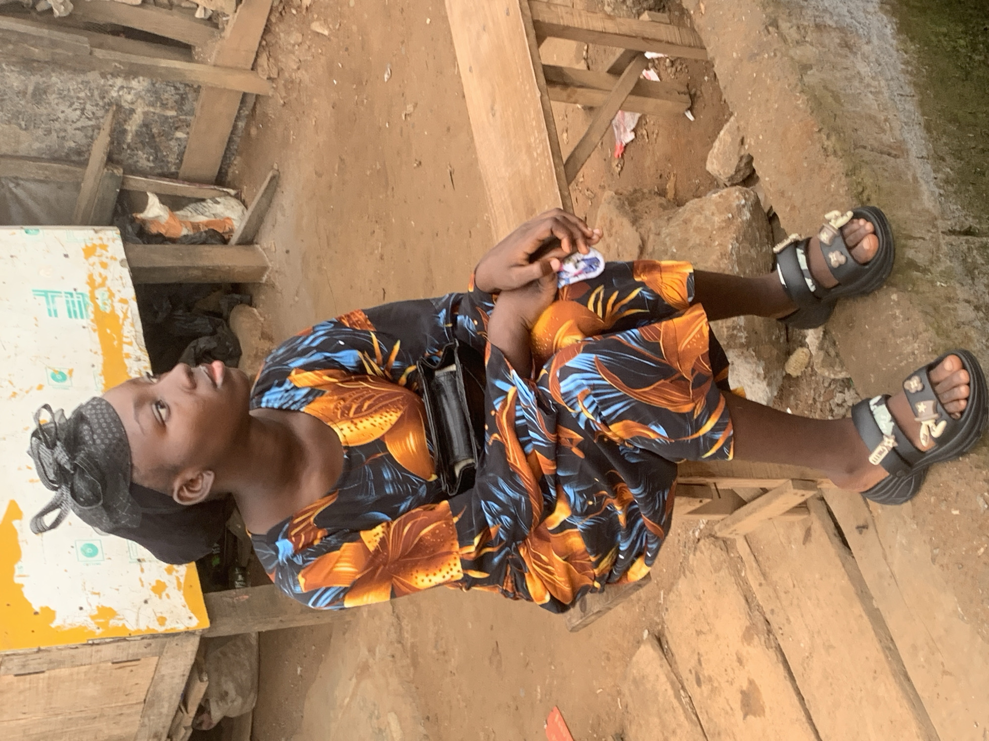 A young Black woman wearing a black headscarf with a decorative fascinator is sitting on a wooden bench in an outdoor market setting in Shagamu, Nigeria. She is looking to her right, with her hands resting on her lap. She is wearing a long-sleeved dress with a vibrant floral pattern of orange, blue, and black. She has a black clutch bag slung across her chest. On her feet, she wears black platform sandals with decorative buckles. The background shows stalls made of wood and corrugated metal, with sacks and debris scattered around, indicative of a bustling, somewhat unkempt market environment. The ground is composed of dirt and rough stones. The lighting suggests it is daytime.