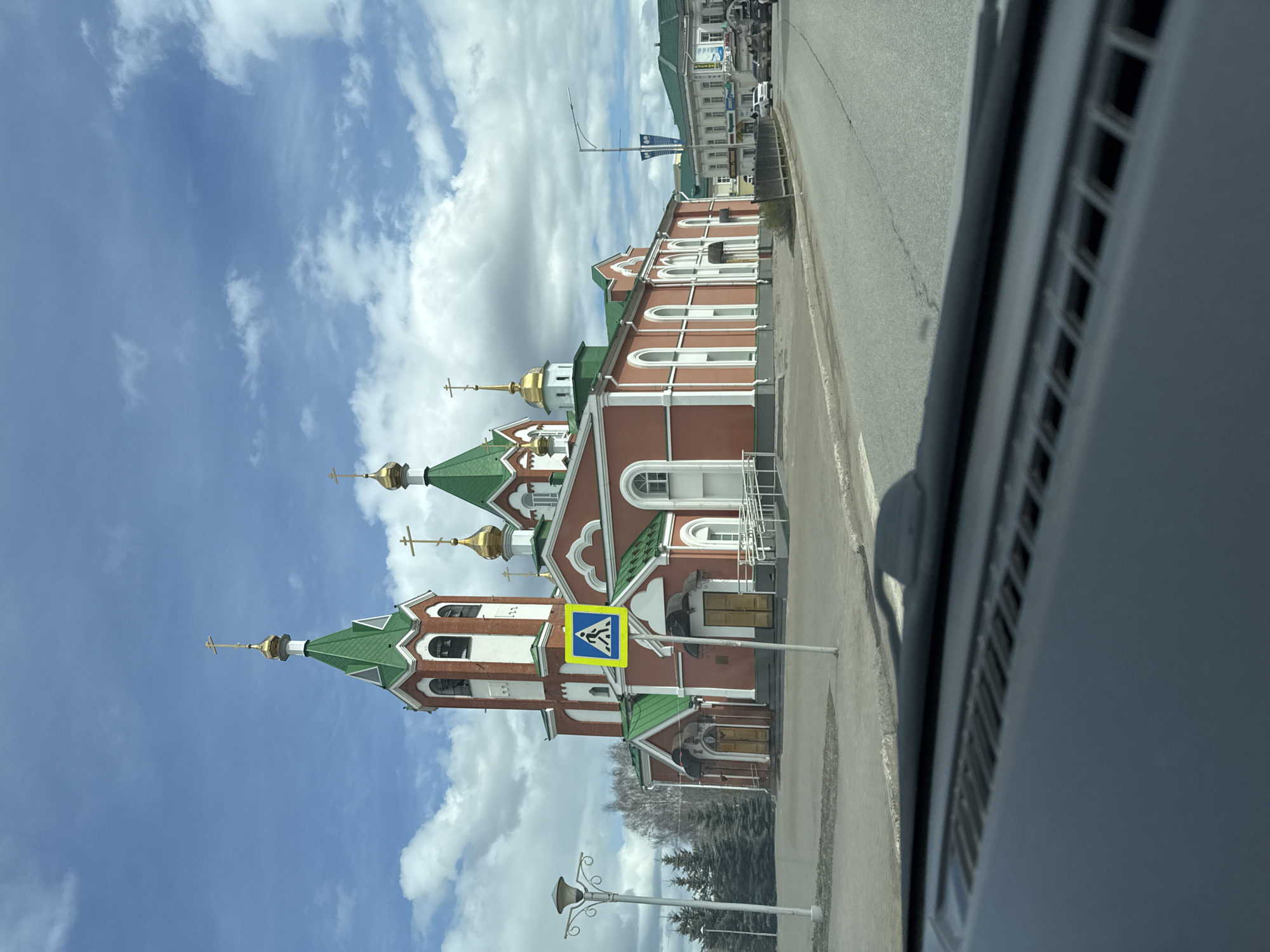 The image captures a vibrant, multi-domed Orthodox church viewed from a slightly elevated perspective, likely from a moving vehicle. The church dominates the left side of the frame, featuring striking red brick walls with white arched window frames and decorative accents. Its numerous roofs are a distinctive green, topped with golden onion domes, each crowned with an Orthodox cross. A prominent yellow square sign with a blue border, indicating a pedestrian crossing, is positioned in front of the church.
To the right of the church, an asphalt road stretches into the distance, suggesting an urban street. The far right side of the image reveals part of a car's interior, possibly the dashboard or A-pillar, partially obscuring the road. In the background, behind the main church, other buildings with lighter facades and green roofs are visible, along with some trees, hinting at a built-up area. A decorative street lamp post stands in the lower left foreground.
The sky is a clear blue adorned with scattered white clouds, indicating daytime with good weather. No specific human activity is captured, the scene portrays a static street view in Glazov, Russia, with the church as the central architectural highlight.