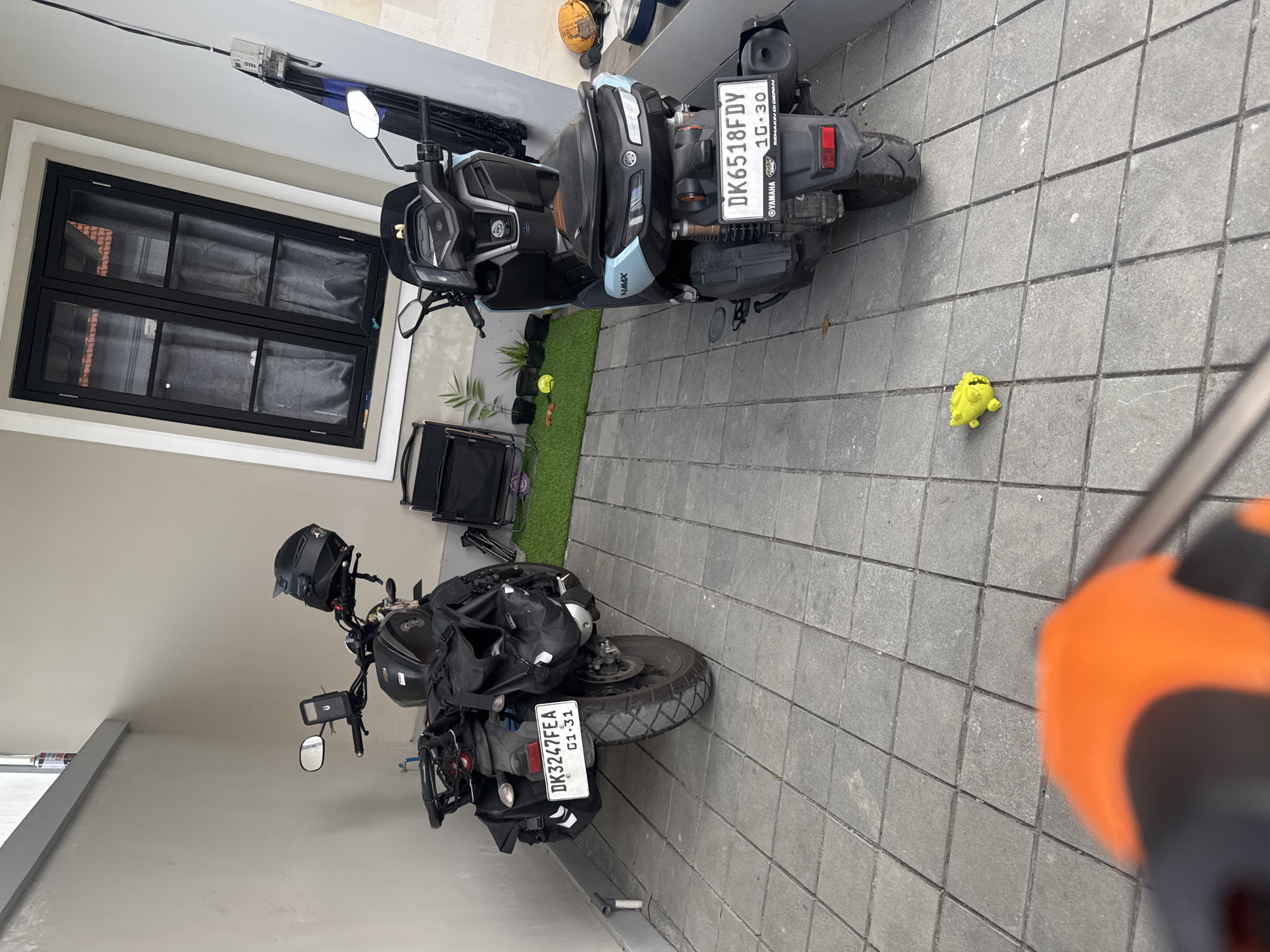 The image depicts an outdoor area with paved stone flooring, likely a driveway or a patio. Two motorcycles are parked side-by-side. The motorcycle on the left is black and appears to be equipped for touring, with saddlebags on either side and a helmet resting on its handlebars. Its license plate reads "DK3247FEA" and "01.31". The motorcycle on the right is a lighter blue and black scooter, with a license plate that reads "DK6518FDY" and "10.30", and has "YAMAHA" branding visible.
In the background, a building with a large black-framed window is visible. To the left of the building, a grey wall extends outwards, and a small, bright yellow toy is on the ground between the two motorcycles. A piece of artificial grass with several small potted plants is also present behind the motorcycles. The overall setting suggests a residential or commercial area within the city of Sukawati, Indonesia. The image is captured from a low angle, with the foreground partially obscured by an orange and black object, possibly a camera or a phone holder.