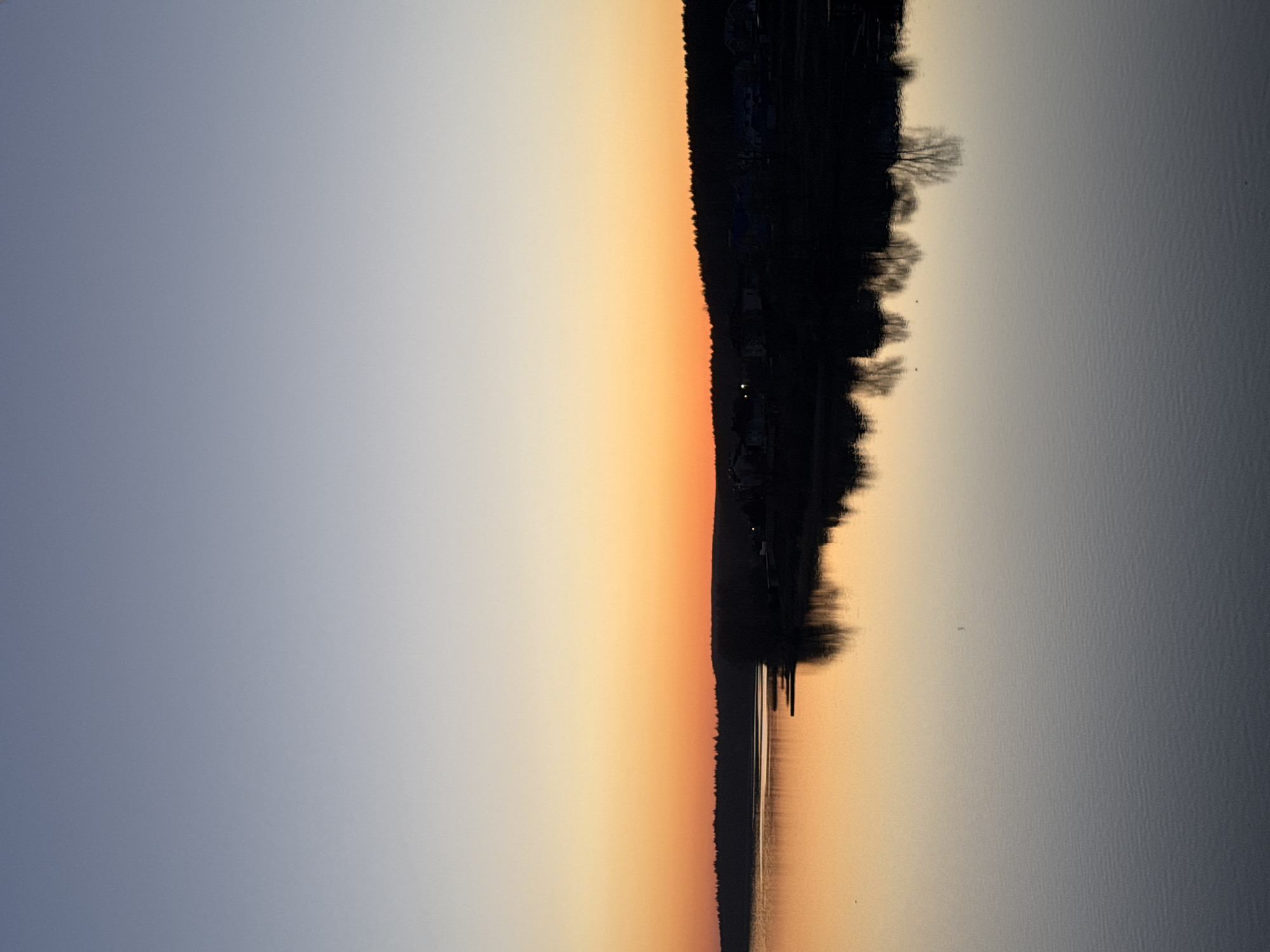 The image shows a sunset over a body of water, likely a lake or a wide river, with a silhouetted shoreline in Votkinsk, Russia. The sky displays a gradient from light blue to orange, reflected in the water. The shoreline has trees and buildings, creating a dark silhouette against the sky. Several pylons are visible on the water. No people are visible.