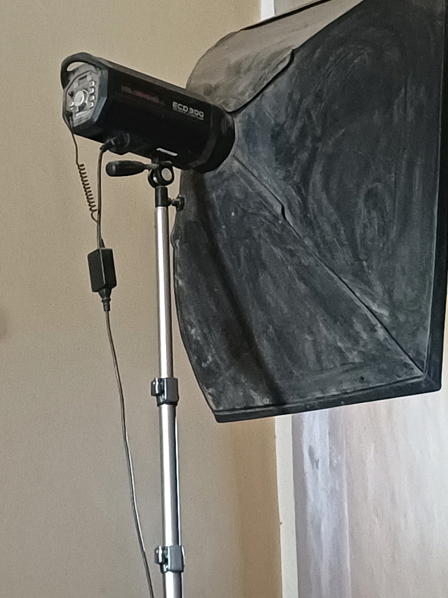 This is a close-up, eye-level, indoor shot of a photography light and softbox. The light is black with the label "ECD 300" on it. It is mounted on a silver metal stand with black clamps. A black cord with a black adapter hangs from the light. A large, black softbox is attached to the light, diffusing the light source. The background consists of a plain, light-colored wall and a white column, suggesting an indoor studio setting. There are no people or animals visible in the image. The lighting is somewhat dim, with some dust visible on the softbox. There is no indication of the specific location within Dumai City, Indonesia.