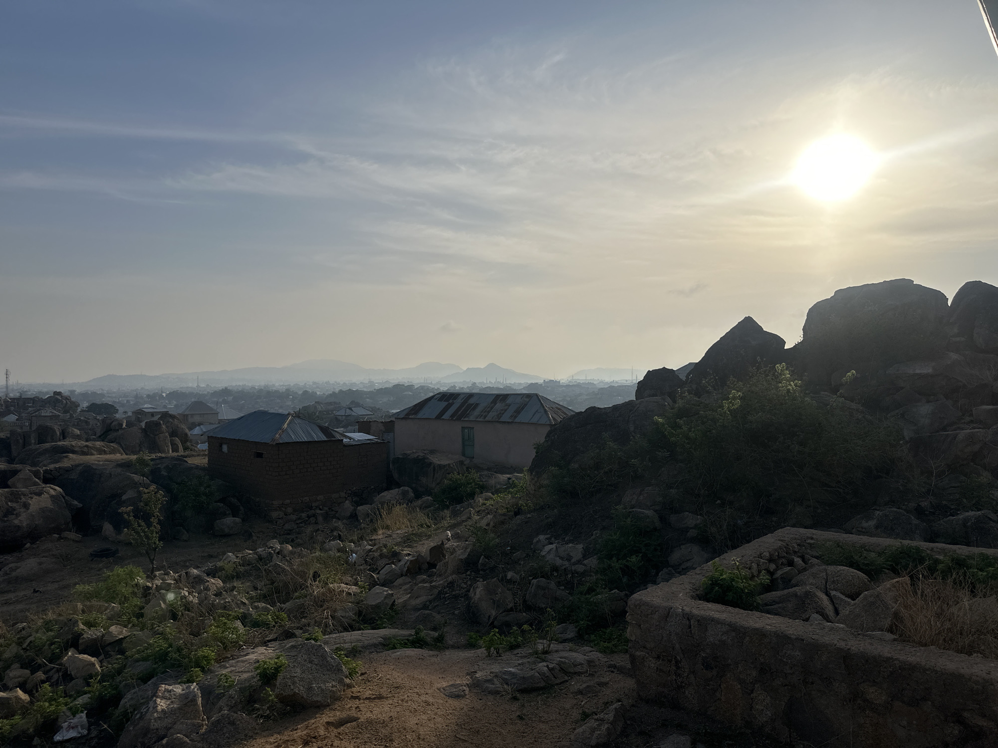 The image depicts a landscape in Jos, Nigeria, likely a rocky elevated area with buildings and vegetation. The sun is high in the sky, casting a hazy light over the scene. In the foreground, there are large boulders, shrubs, and what appears to be a stone-lined structure. Two buildings with corrugated metal roofs are visible among the rocks. In the background, there is a cityscape with more buildings and distant mountains visible through the haze. The environment is rocky and uneven. No people are visible in the image.