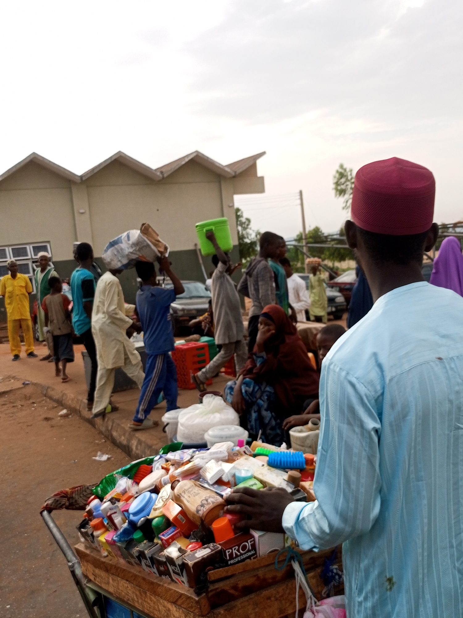 The image depicts a busy street scene in Gombe, Nigeria, with a crowd of people gathered around a cart laden with various goods. In the foreground, a man wearing a light blue kaftan and a maroon cap faces away from the camera, his hands resting on the cart. The cart itself is overflowing with an assortment of items, including bottles of cleaning supplies, soaps, and boxes of what appear to be toiletries.
Several individuals are visible in the midground, some carrying bundles on their heads or arms. One person in a blue t-shirt and striped pants is walking, while another in a cream-colored garment is seen from the side. To the left, a man in a bright yellow outfit stands, and further back, a woman in a burgundy hijab is seated. The background shows a light-colored building with a distinct pitched roofline, likely a commercial or institutional structure. Cars are parked in the distance, suggesting a public area. The ground is unpaved, indicating a typical roadside market environment. The overall atmosphere is one of commerce and daily activity in a Nigerian city.