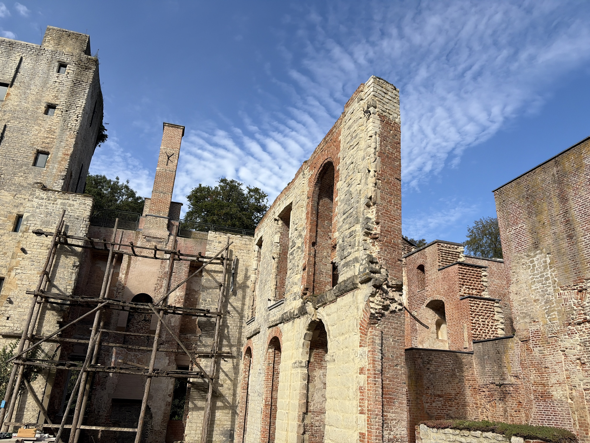 This is a low-angle, eye-level, full shot of the ruins of the Abbey of Grimbergen in Grimbergen, Belgium. The ruins are constructed of red brick and pale stone, and are weathered and aged. Scaffolding made of wooden poles is erected on the left side of the image. The sky is a clear blue with scattered white clouds. The image is taken on a sunny day. There are no people visible in the image.