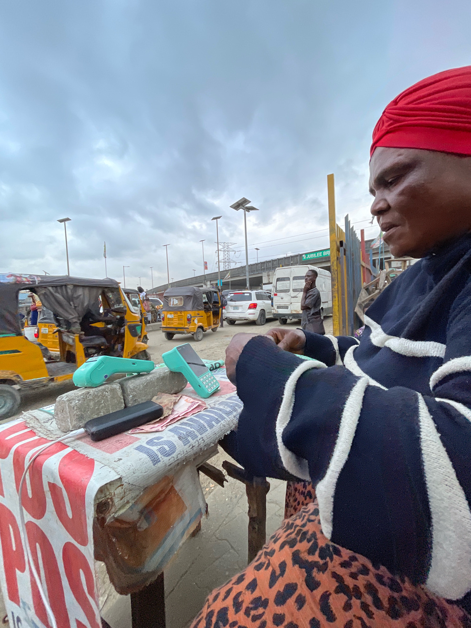 The image depicts an outdoor scene in Ajah, Nigeria, likely near a transportation hub or market, given the presence of several yellow auto-rickshaws (keke-napep). The sky is overcast with heavy clouds. In the foreground, a woman wearing a red headwrap and a dark sweater with white stripes is seated and looking down at a table. Her lower body is covered by a patterned skirt. On the table, which is draped with a white and red-patterned cloth and appears to be made of a rough material, are several items including two light green point-of-sale (POS) devices, two concrete blocks, some currency notes, and a black power bank with a white cable.
In the middle ground, various auto-rickshaws are visible. Some are stationary, with occupants inside or standing nearby, while others appear to be in motion. In the background, a white van and a white SUV are on a road, suggesting a street or thoroughfare. A man is standing by the white van, seemingly engaged in a conversation or looking at something. Beyond the road, there's an elevated structure that appears to be a bridge or flyover, with the text "JUBILEE BR" visible on a sign attached to it. The overall environment suggests a busy urban setting with ongoing commercial and transportation activities.