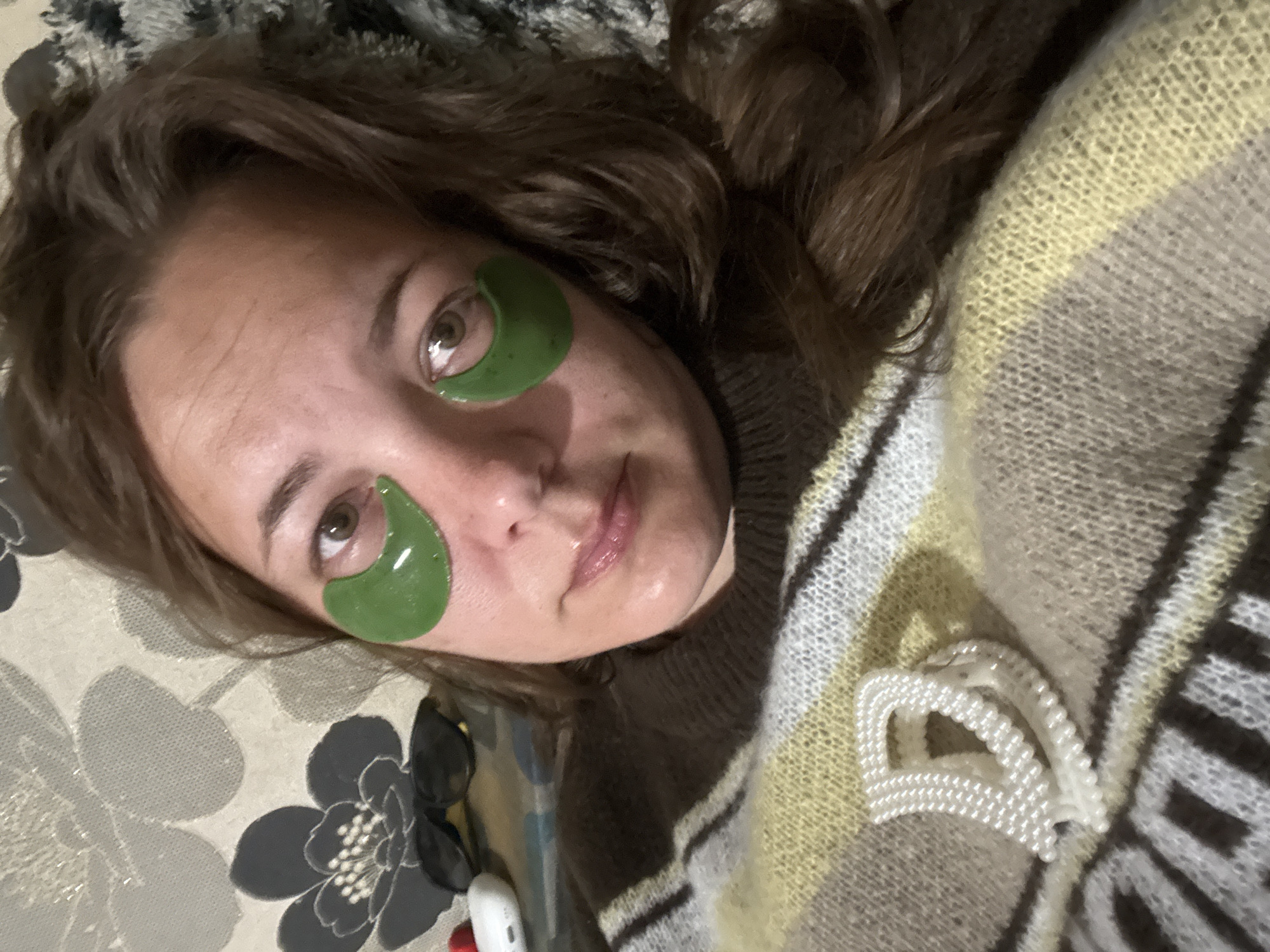 A person is lying down with green under-eye patches on. They have light skin and brown hair. They appear to be wearing a brown sweater or shirt, and a striped shawl or blanket with off-white, gray, and brown stripes. A white pearl beaded hair clip is visible on the shawl. The person is on a floral patterned surface. The location is not discernible from the image.