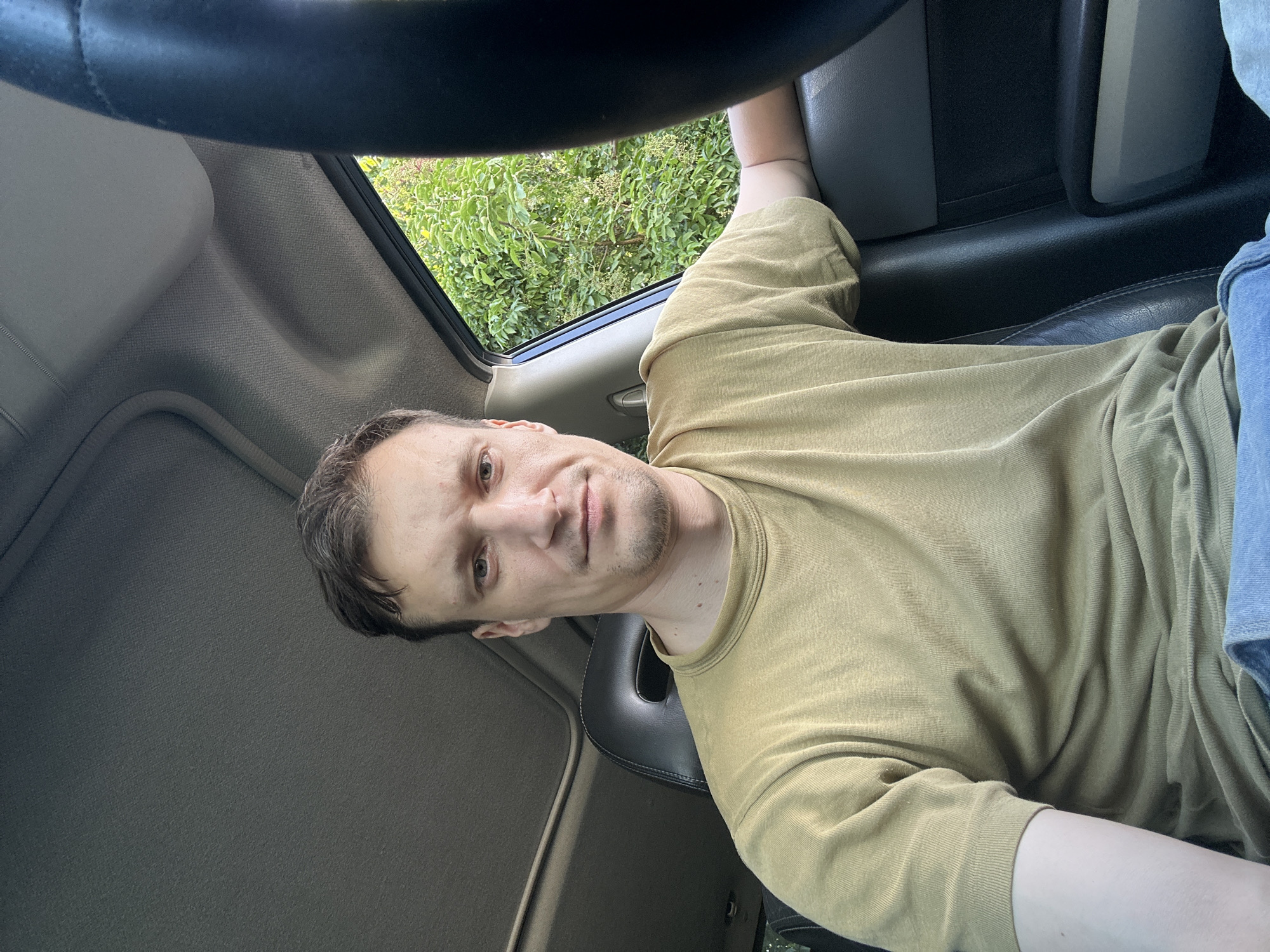 A man is sitting in the driver's seat of a car. The man has short, dark brown hair and is wearing a light brown t-shirt and blue jeans. He is looking at the camera and has a neutral expression. The car's interior is visible, including the dashboard, steering wheel, and seats. Outside the car, lush green trees and foliage can be seen. The scene appears to be a sunny day. The image is a selfie taken by the man in the car. The location is the city of Shatava, Ukraine.