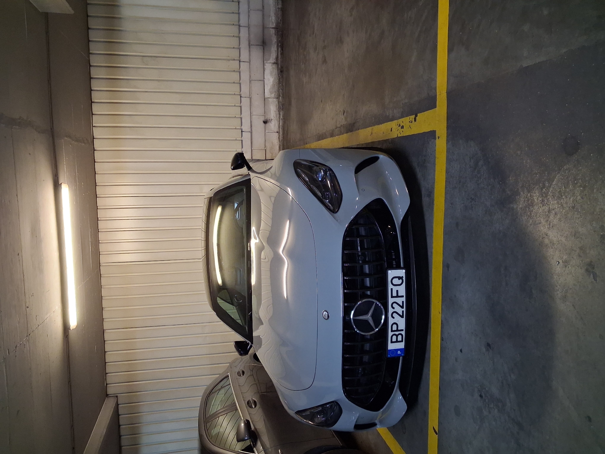 A parked, light-gray Mercedes-Benz AMG GT (likely the Black Series variant) is visible in an indoor parking garage. The license plate reads "BP 22FQ," a Polish license plate. The car is parked next to a yellow painted line, delineating a parking spot. To the left of the car is a wall with horizontal, corrugated metal cladding, illuminated by a fluorescent light fixture. Another vehicle, darker in color, is visible partially obscured behind the AMG. The garage floor is concrete. The image shows no people, and no actions or interactions are taking place. It's impossible to determine the precise location within Lisbon without further information.