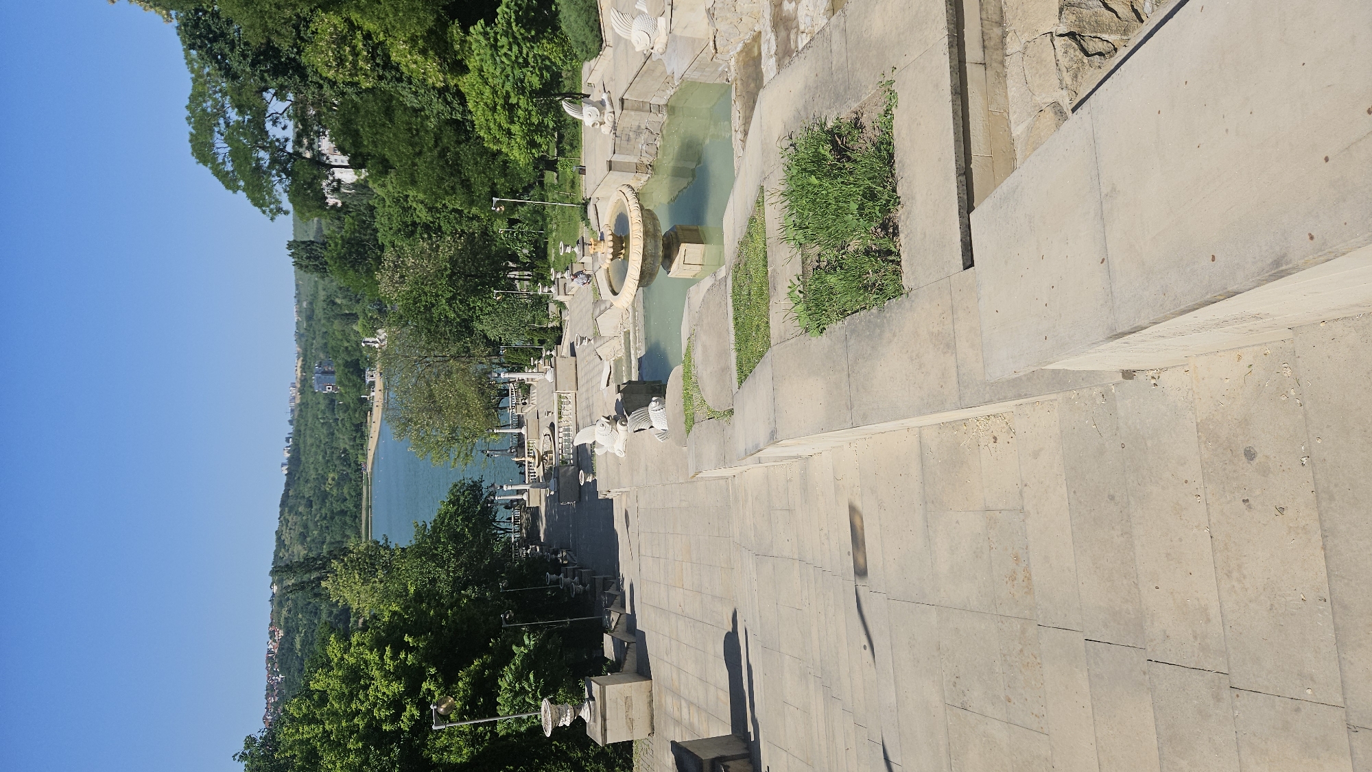 This image depicts a view of the Cascade Stairs in Chișinău, Moldova, featuring a cascading fountain system with multiple levels. The stairs are constructed of light-colored stone and extend downwards toward a body of water, likely a lake or river. Lush green trees are visible along the sides, lining the pathway leading down the stairs. A bright blue sky is present in the background. There are no visible people in this image.