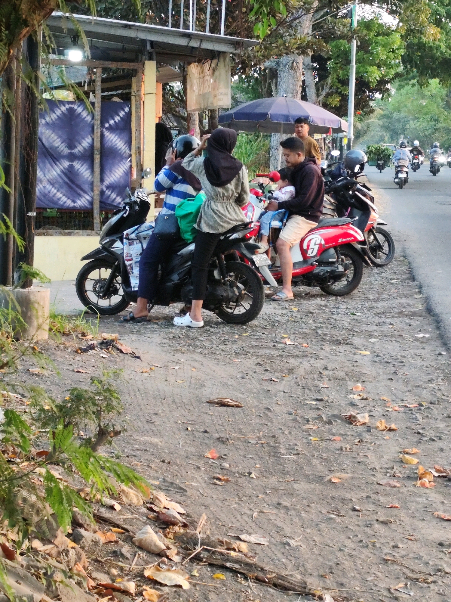 The image depicts a roadside scene in Bantul, Indonesia, with several people on motorcycles. In the foreground, two individuals are seated on a black scooter. The person in the back, wearing a black hijab and a light-colored patterned jacket, is adjusting a helmet. The person in front, wearing a blue and white striped shirt and dark pants, has a green bag on their shoulder and is wearing sandals. They are positioned near a stall with a blue and white tie-dye fabric hanging as a partition.
To the right of the black scooter, a red and white scooter is parked. A man in a dark hoodie and beige shorts is holding a baby in a pink and white outfit. Another person, partially visible and wearing a brown shirt, stands behind them under a dark blue umbrella.
In the background, a road is visible with more people on motorcycles, some carrying bundles of greenery. The environment appears to be a street lined with trees and some vegetation, with dry leaves scattered on the ground. The lighting suggests it is daytime, possibly late afternoon. The overall scene conveys a sense of everyday life and transportation in a semi-urban or rural setting in Bantul.