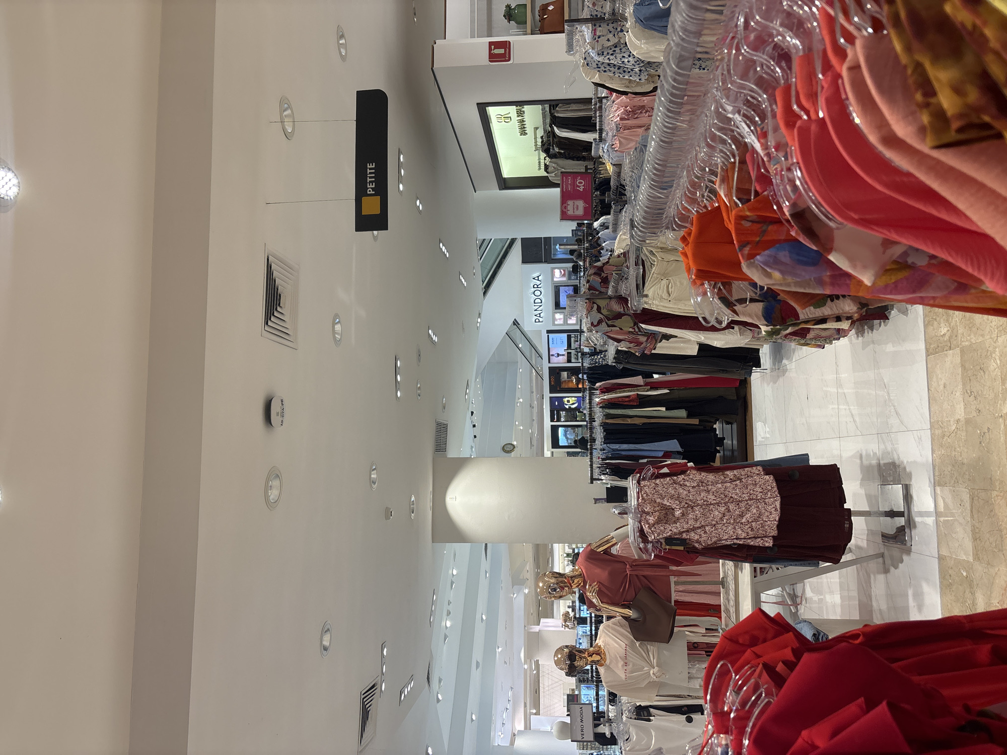 The image shows the interior of a clothing store, likely a department store, in what appears to be Boca del Río, Mexico. Racks of clothing on hangers are visible, with a mix of colors and styles on display. A sign indicating "PETITE" hangs from the ceiling. A "PANDORA" display is seen in the distance. Mannequins are present, displaying clothing. The environment is brightly lit with recessed ceiling lights. The floor is tiled. There are no people immediately visible in clear focus, though there are figures in the background near the mannequins.