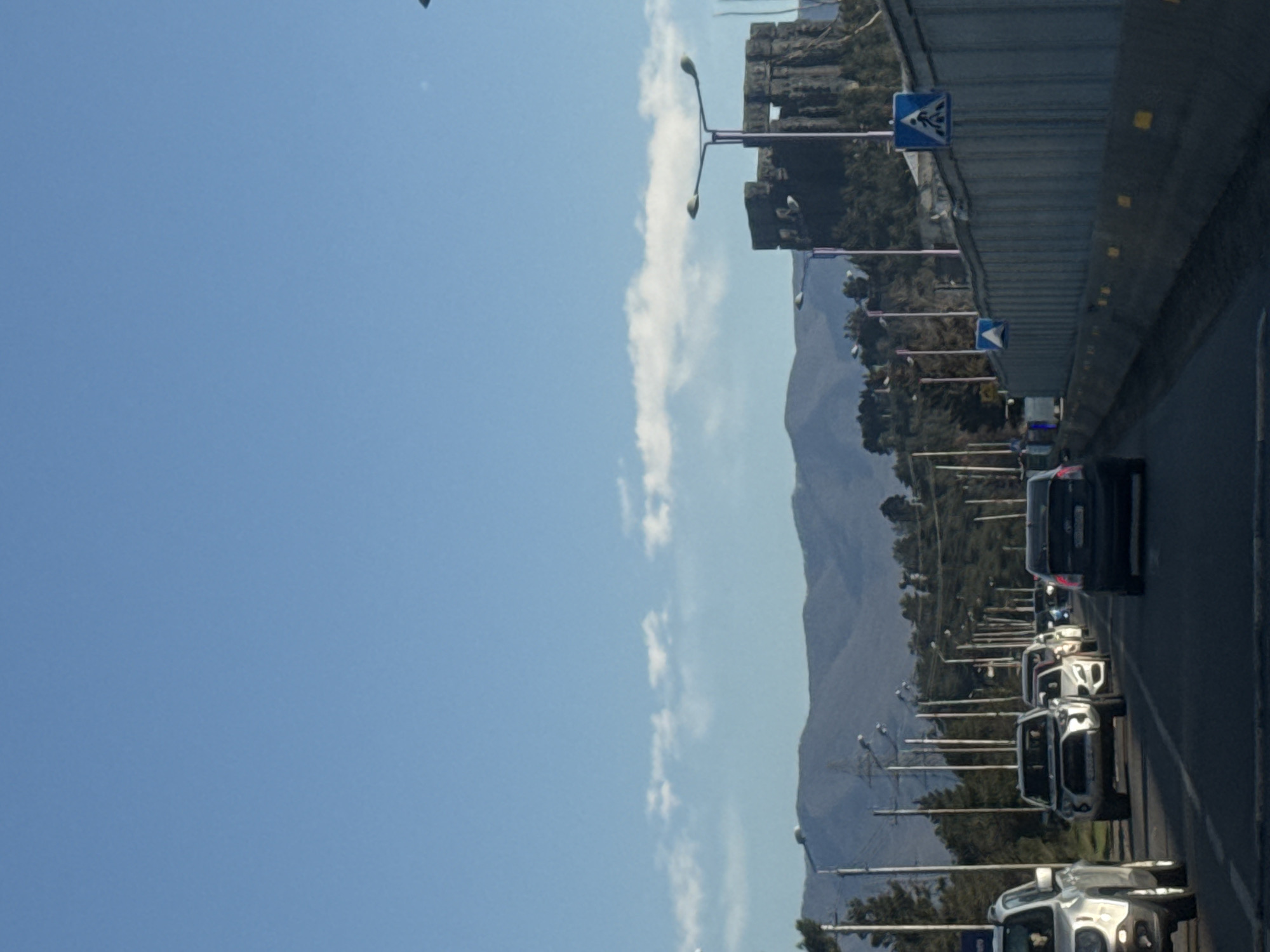 View from the highway in Tbilisi, Georgia, of a line of cars stopped in traffic. To the left, a clear blue sky with a line of fluffy white clouds. A mountain range parallels the highway. Lampposts line the highway. Road signs are overhead.