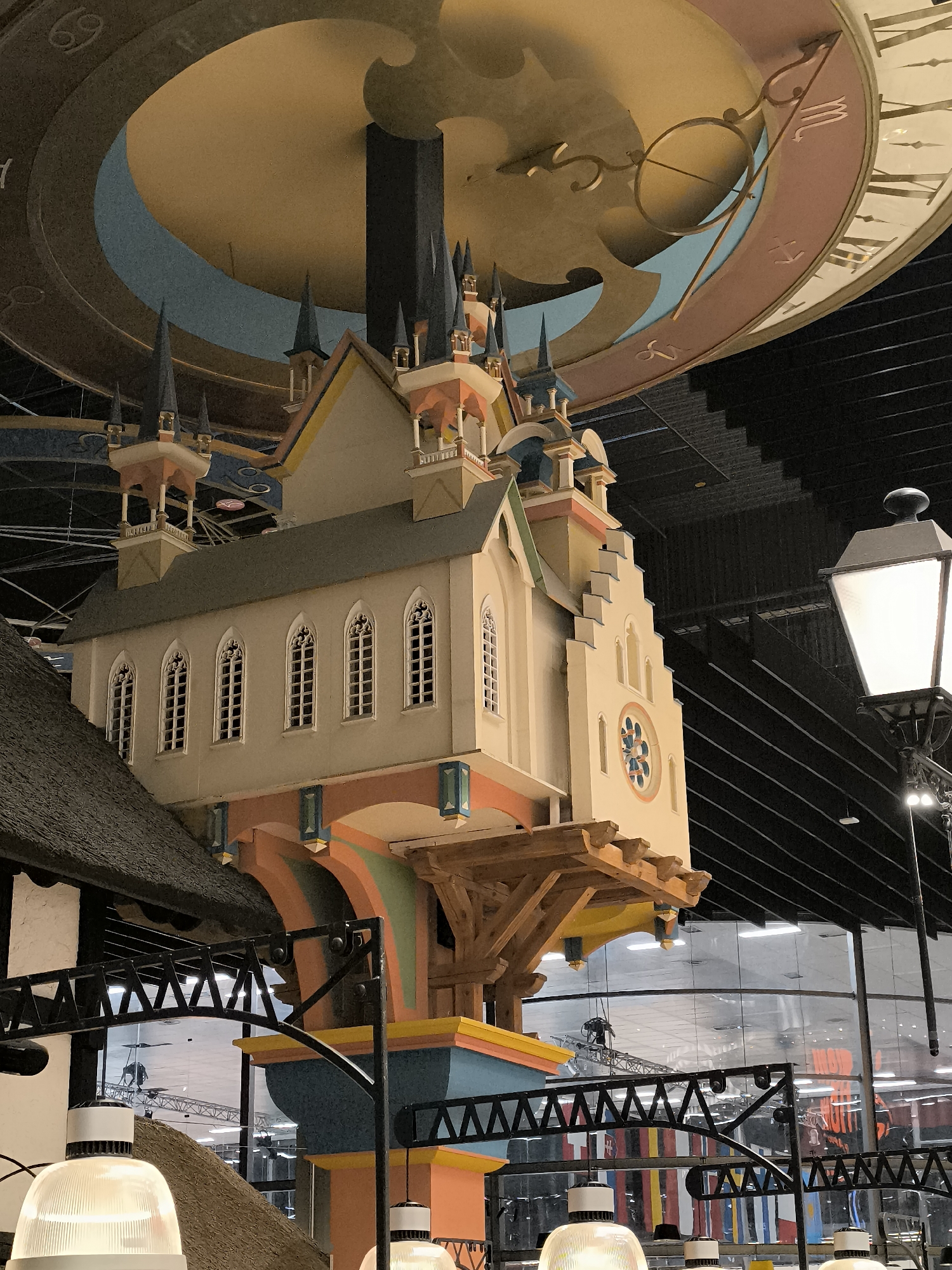 This is an indoor, eye-level, medium shot of an ornate, multi-story clock tower structure suspended from the ceiling, with a large astrological clock face visible above. The clock tower is painted in pastel colors with architectural details resembling a church or castle, including spires, arched windows, and decorative supports. A street lamp is positioned to the right of the clock tower, emitting light. The background consists of a dark, ribbed ceiling and a large, reflective glass wall revealing glimpses of a larger interior space with industrial lighting and various banners or flags. This structure is located at the Klaipėda Clock Museum (Laikrodžių muziejus) in Klaipėda, Lithuania, a unique museum dedicated to the history and artistry of clocks and timekeeping.