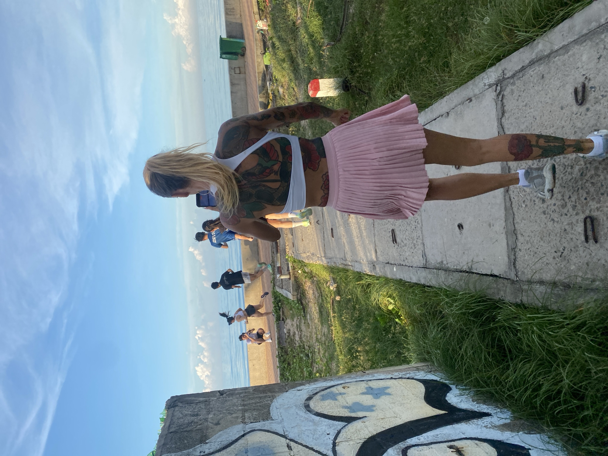 A woman with extensive tattoos on her back and legs, wearing a white sports bra and a pink pleated skirt, is captured from behind as she walks on a concrete path. She is holding a smartphone in her right hand. The path leads towards the sea under a blue sky with scattered clouds. In the background, several individuals are visible running or walking along a boardwalk adjacent to the water. A concrete wall with graffiti is on the left side of the frame, and overgrown grass borders the path on the right. Two green trash bins are positioned near the water's edge. The scene appears to be set in a public outdoor area, possibly a coastal promenade in Phu Thuy Ward, Vietnam, given the context provided.