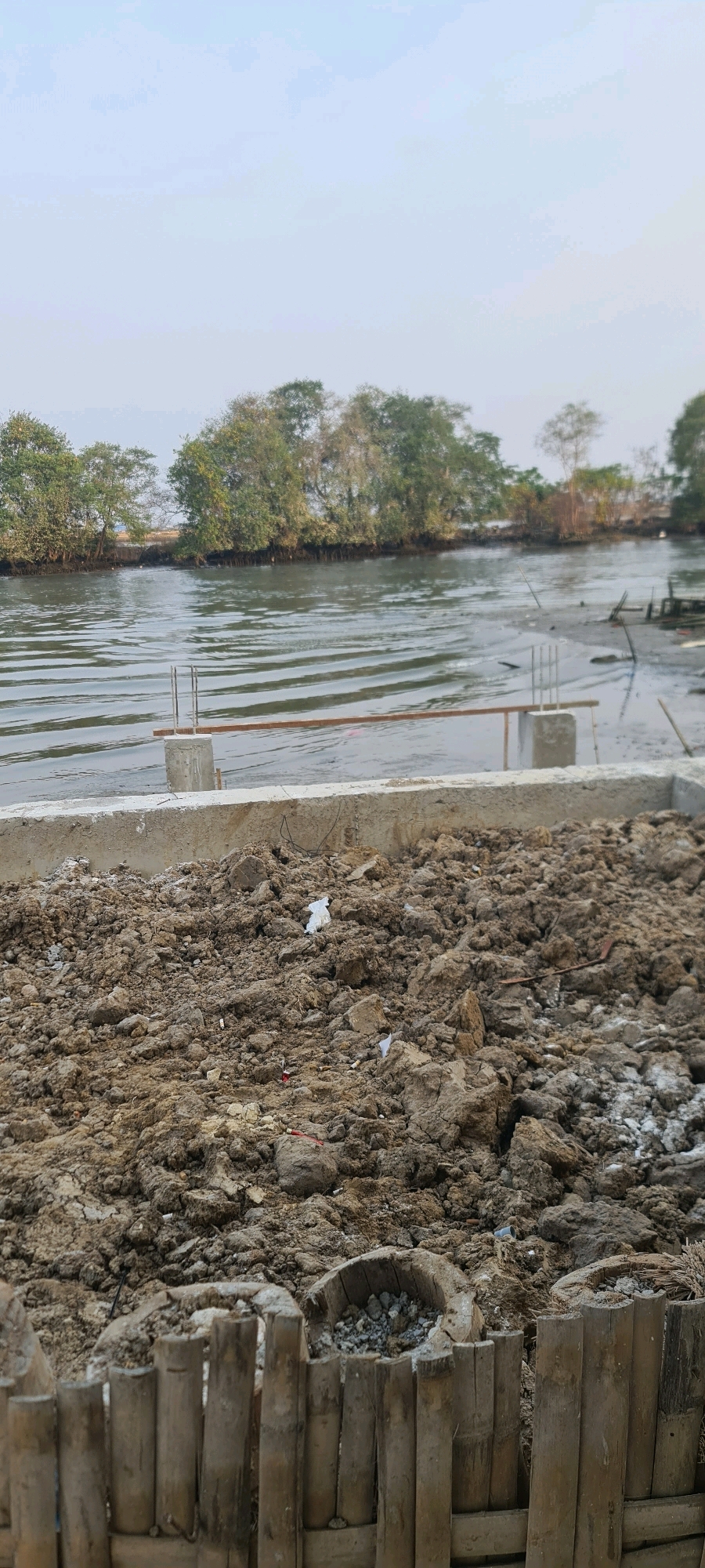 The image shows a construction site near a body of water, likely a river or canal, in Juwana, Indonesia. In the foreground, there is a pile of dark, damp soil, appearing to be excavated earth, with some scattered debris like white paper and what might be cigarette butts. A low concrete barrier runs along the edge of the excavated area, with exposed rebar protruding from a concrete pillar. Behind the barrier, the water is rippled and appears shallow near the shore, revealing mudflats.
Across the water, a line of lush green trees forms a dense canopy along the distant bank. The sky above is a pale blue with some hazy clouds. The overall impression is of a developing construction project in a natural, possibly coastal or riparian, environment. The scene is static, with no people visible, suggesting a pause in construction activity. The bamboo fencing in the immediate foreground adds a rustic element to the otherwise utilitarian construction scene.