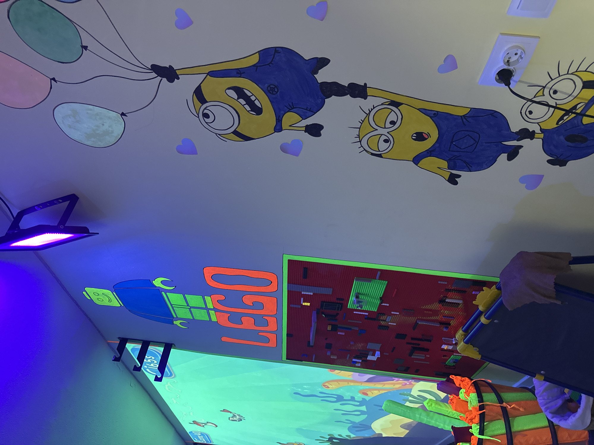 The image depicts an indoor scene, likely a child's room or play area, illuminated by blacklights that cause fluorescent elements to glow. On the wall, a large drawing of a LEGO figure with a green head and body and blue overalls stands prominently. To the right, a mural features two Minions from the Despicable Me franchise holding onto balloons, with smaller purple hearts scattered around them. Below the LEGO figure, the word "LEGO" is written in large, orange letters with a fluorescent outline. Further down, a red board filled with various LEGO bricks serves as a display or building surface. To the left of the LEGO display, a projection of an underwater scene with fish and coral is visible on the wall, with the text "Score-40" and "1:65s" displayed on it. In the foreground, a bucket filled with brightly colored, fluorescent green and orange cylindrical objects, possibly pool noodles or similar toys, is visible. A dark blue easel with a cloth draped over it is positioned in front of the LEGO display. An electrical outlet with two plugs and a black cord extending from it is mounted on the wall near the Minion mural. The overall environment is colorful and playful, with a strong emphasis on LEGO and popular animated characters.