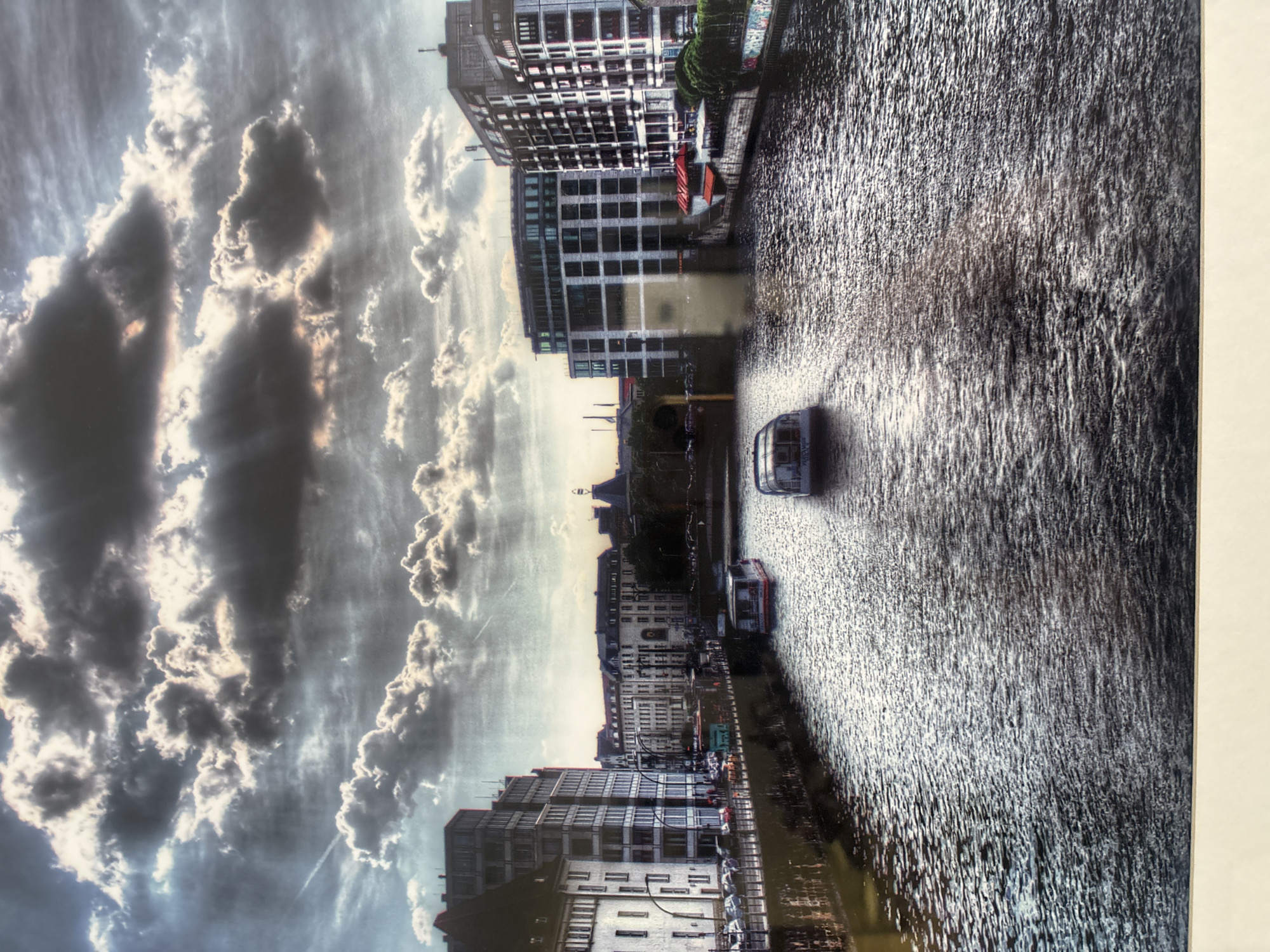 The image depicts a canal scene in a European city, possibly Berlin, Germany, given the architectural style of the buildings lining the waterway. The sky is dramatic with heavy, textured clouds, through which rays of sunlight are breaking. Two tour boats are navigating the water, leaving wakes behind them. The buildings on either side are a mix of modern and older architecture, with apartments, offices, and some businesses with outdoor seating visible. The canal is a dark, reflective surface, mirroring the sky and the surrounding structures. There are no discernible people in the immediate foreground, though there appears to be activity on the canal banks in the distance, with cars and pedestrians. The overall impression is one of urban life along a historic waterway under a dynamic sky. The image is NOT located in Dubai, United Arab Emirates.
