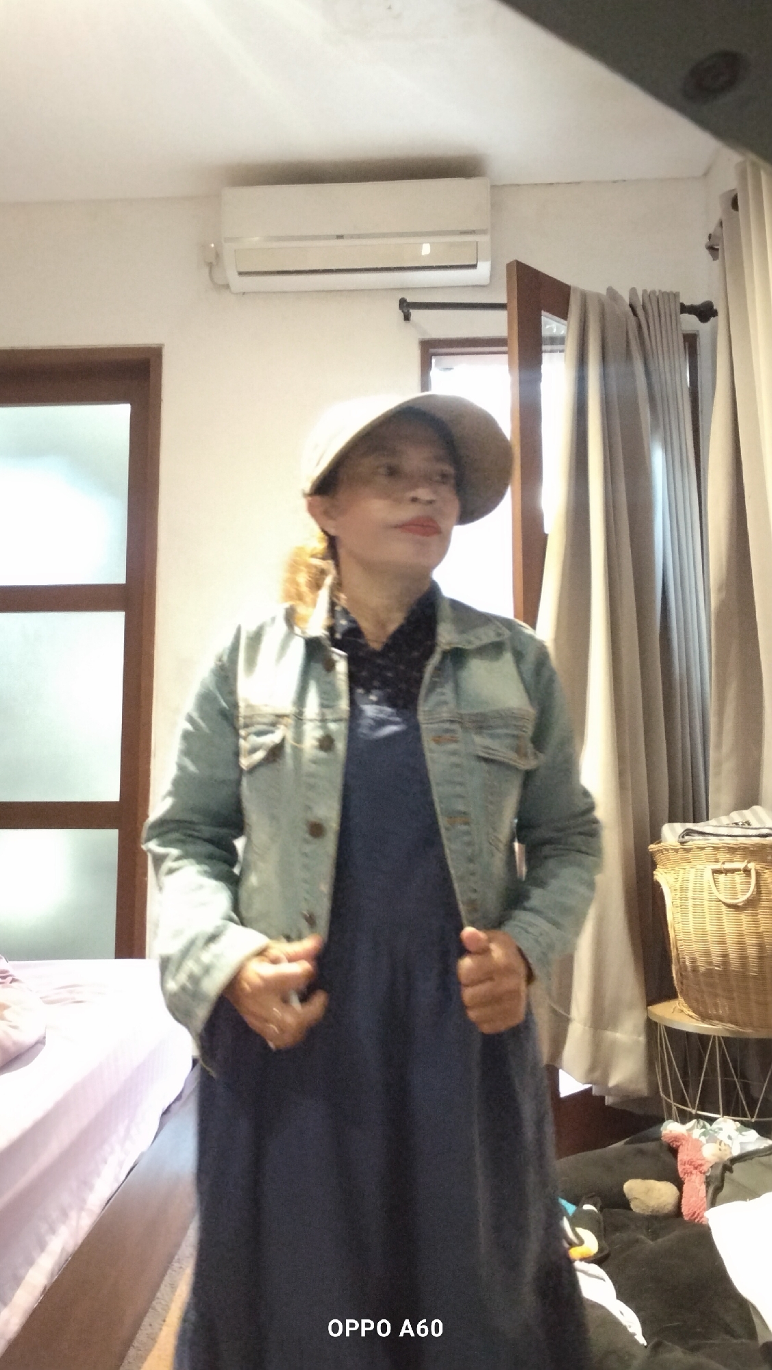The image shows a woman standing in a room in North Kuta, Indonesia. She is wearing a denim jacket over a dark blue dress and a light-colored cap. She is holding the front of her jacket with both hands. In the background, there is an air conditioning unit mounted on the wall, a window with wooden frames and sheer curtains, and a wicker basket on a small table. To the left of the woman, a bed with a light purple cover is partially visible. The floor appears to be dark. The room has a generally bright and airy feel.