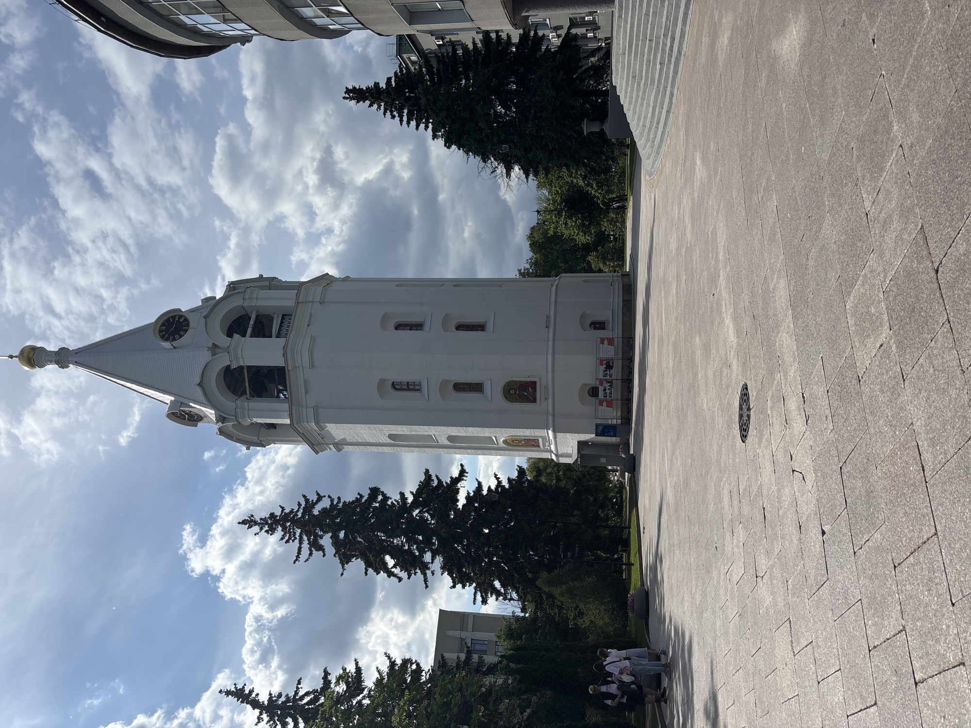 This image depicts a sunny outdoor scene, likely a public square or park, featuring a tall, white bell tower or church with a prominent golden dome and a clock face. The building, consistent with Russian Orthodox architecture, stands on the left side of the frame. Its upper section features arched openings for bells, and its lower parts display small windows and colorful religious mosaics or icons embedded in the wall.
The sky above is bright blue with scattered white, cumulus clouds, indicating a clear, sunny day, likely mid-day or early afternoon. Tall, dark green coniferous trees frame the white tower and extend into various parts of the scene, providing shade and natural elements.
The foreground and much of the right side of the image are covered by a light-colored, meticulously paved surface made of rectangular stone or concrete blocks. A circular metal grate is visible in the pavement towards the right. On the far right, a set of broad, light-colored steps ascend upwards, suggesting a multi-level public space.
A small group of approximately three to four people, casually dressed, are visible on the far right, gathered on the paved area near the trees. They appear to be resting or conversing. In the upper left background, parts of modern-looking buildings with windows are visible, contrasting with the historical tower.
No legible text is visible on any signs or objects within the frame. The scene conveys a peaceful urban environment in Nizhny Novgorod, Russia.