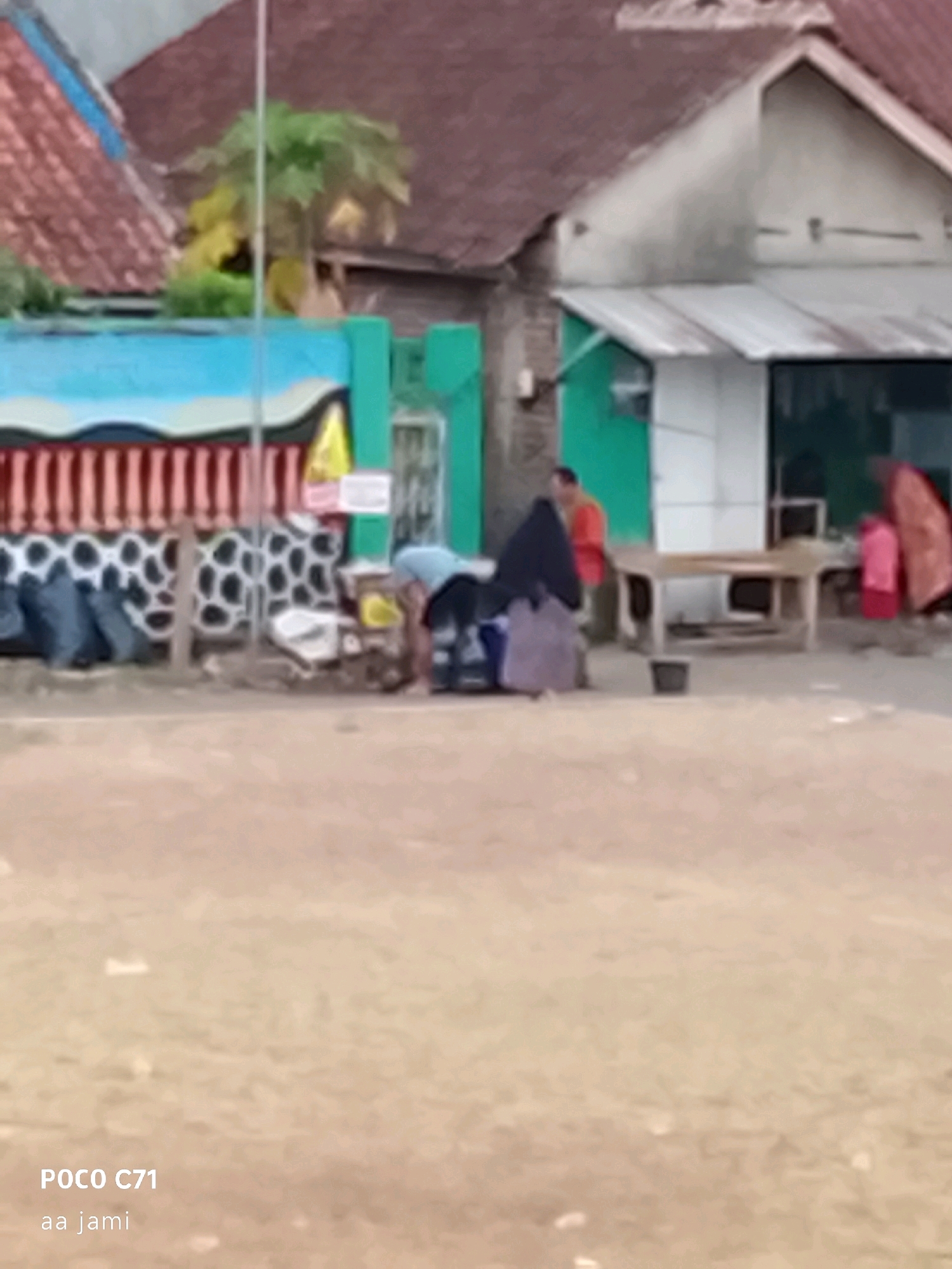 The image depicts an outdoor scene with a focus on two individuals and their surroundings. In the foreground, a person wearing a light blue shirt and dark pants is bending over, appearing to be engaged in some activity on the ground. To the right and slightly behind this individual, another person is partially visible, wearing an orange shirt and standing. In the background, a building with a reddish-brown roof is prominent, with a white section and a green door. A table-like structure is positioned in front of the building. Several dark, possibly garbage bags, are lined up on the left side of the image, near a fence with a black and white spotted pattern. The ground is a sandy or dirt surface. The overall impression is a casual, possibly domestic or commercial setting. The text "POCO C71 aa jami" is overlaid in the bottom left corner, suggesting the image was taken with a POCO C71 phone.