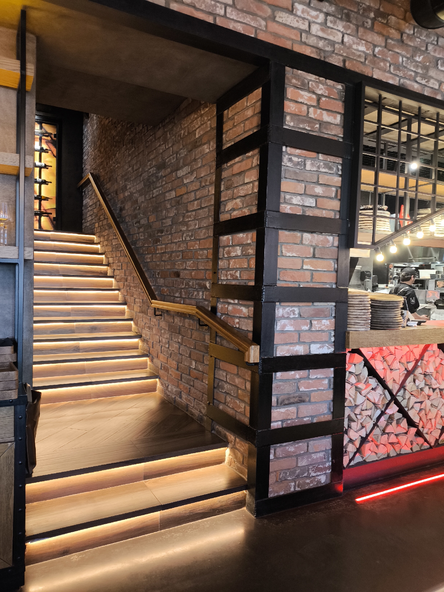 The image depicts an interior scene, likely a restaurant or bar in Izhevsk, Russia, though specific street or establishment identification is not visually present. The perspective is from a slightly elevated position, looking into a multi-level space.
The dominant features are a brightly lit wooden staircase on the left and a partially visible kitchen/bar area on the right.
**Staircase:**
An ascending staircase occupies the left and central portion of the frame. It features light-colored wooden treads with darker risers. Each step is equipped with a linear warm-toned LED strip light positioned beneath the tread, casting an upward glow. A wooden handrail, supported by dark metal brackets, runs along the right side of the staircase, affixed to an exposed brick wall. At the top of the stairs, a dark alcove reveals a vertically oriented wine rack or display, illuminated from behind with a warm light, showcasing multiple wine bottles.
**Structural Elements:**
The interior design incorporates an industrial aesthetic. Walls are constructed from exposed red-brown brickwork with white mortar. Dark, possibly black, metal beams and vertical supports frame sections of the brickwork and act as structural or decorative elements, particularly evident in a prominent column-like structure in the mid-ground.
**Kitchen/Bar Area (Right):**
To the right, behind the brick column, a segment of a commercial kitchen or bar is visible. A light-colored wooden counter extends into the frame. On this counter, several stacks of round wooden boards, possibly pizza peels, are arranged. Behind the counter, one individual is observable. This person is wearing a dark uniform, including a dark cap, and appears to be engaged in work, facing away from the camera. Stainless steel kitchen equipment is discernible in the background. Overhead, multiple bare bulb pendant lights provide illumination.
The base of the counter below the wooden top is clad with neatly stacked firewood, arranged in a grid pattern, and illuminated by a reddish-orange light from within or behind. A horizontal red neon strip light runs along the floor directly in front of this firewood feature.
**Flooring:**
The visible floor at the bottom of the image is a dark, smooth surface, possibly polished concrete or large-format tiles.
**Lighting and Atmosphere:**
The scene is well-lit, primarily by the warm LED strips on the stairs and the pendant lights in the kitchen area. The overall color palette is warm, dominated by the brick, wood, and artificial lighting, creating an inviting ambiance. No other individuals are clearly visible or identifiable within the frame.